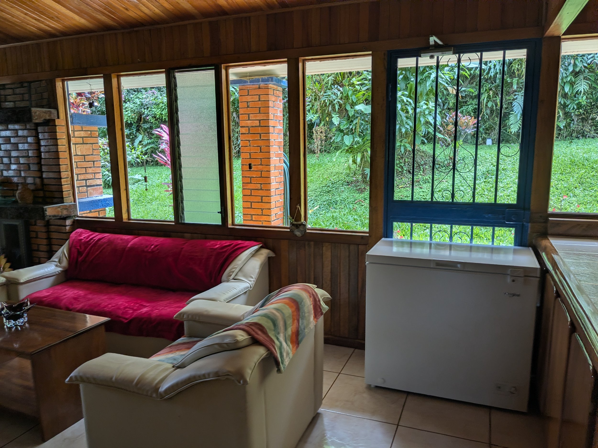 Living room with wooden walls and large windows looking out to greenery, a white refrigerator, a white sofa with red and striped throws, and a wooden coffee table.
