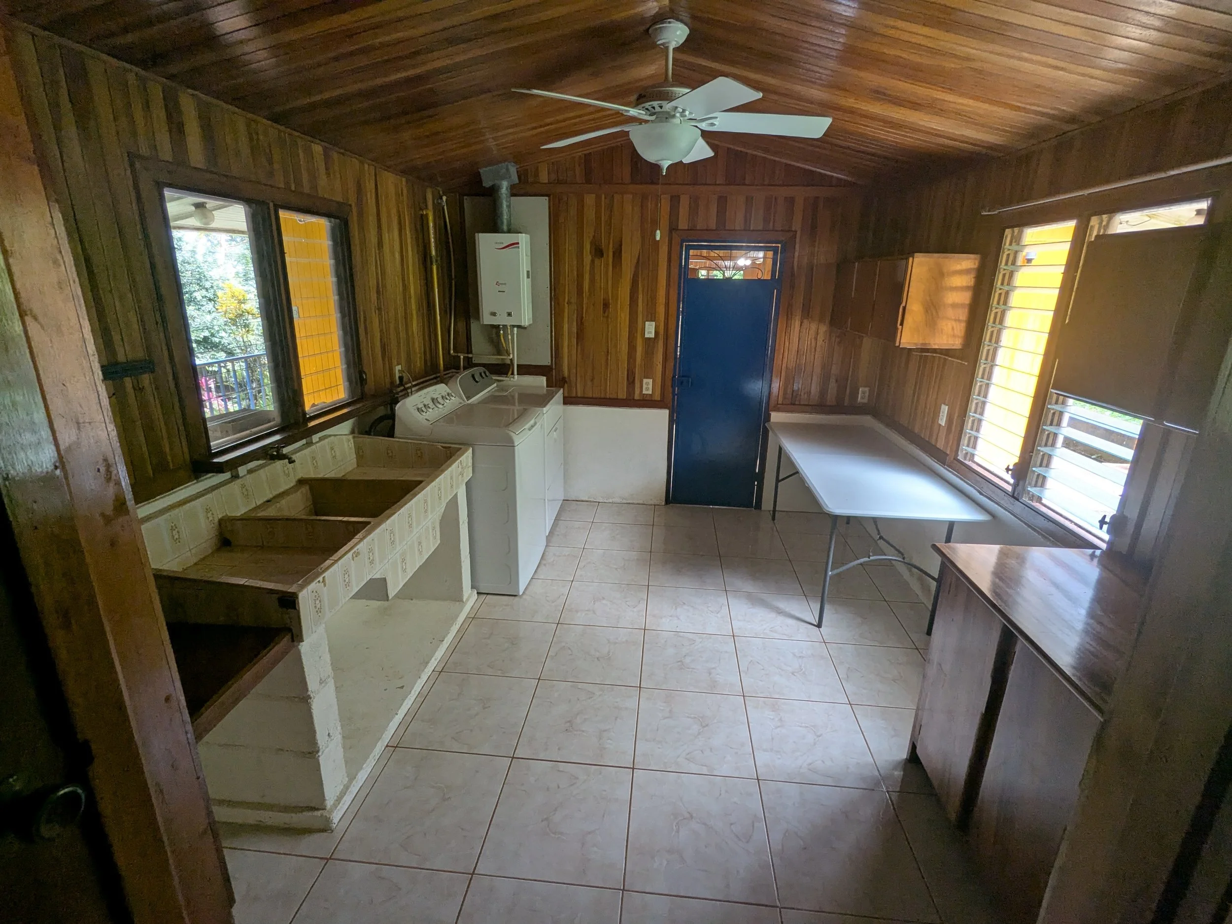 A laundry room with wood-paneled walls and ceiling, a ceiling fan, windows with wooden shutters, a washing machine, a utility sink, a small overhead water heater, a blue door, a folding table, and a wooden cabinet.