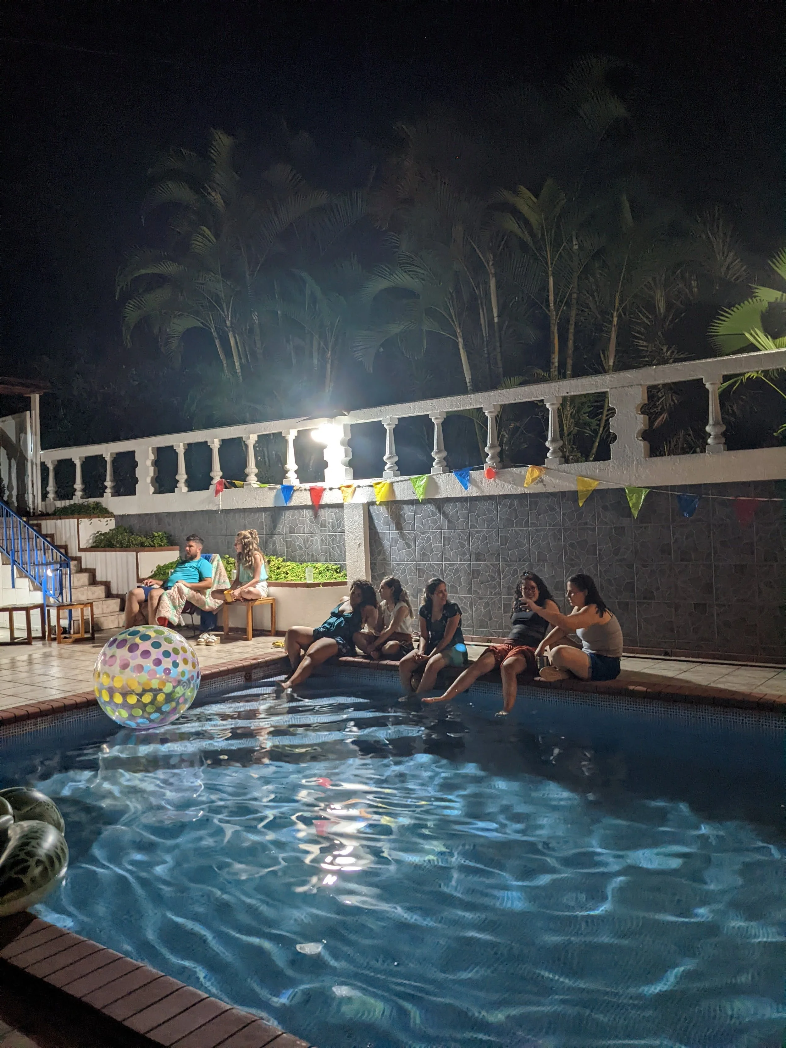 People sitting by a swimming pool at night, some with feet in water, others sitting on the edge, with colorful bunting hanging above, surrounded by palm trees.