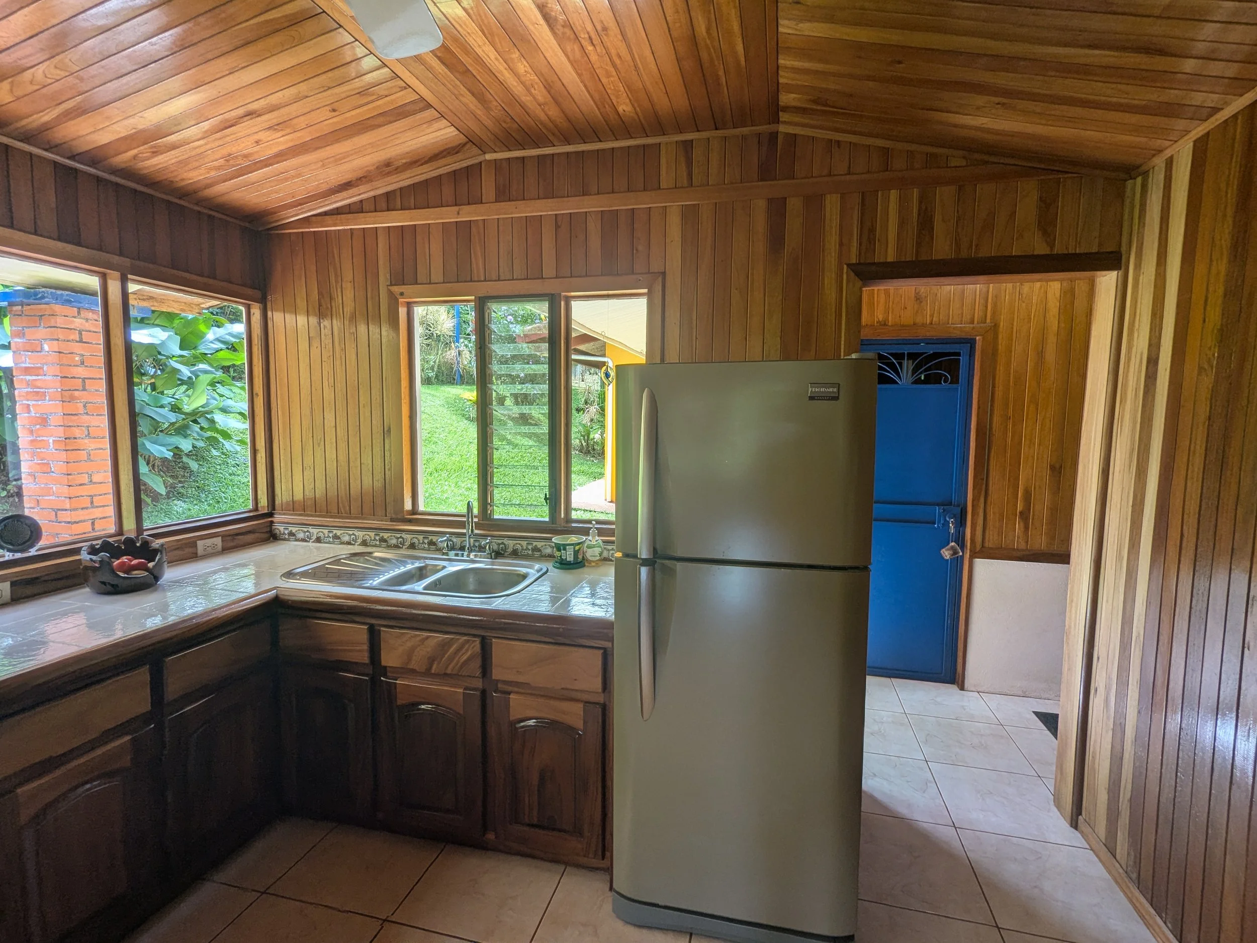 Kitchen with wooden paneled walls and ceiling, stainless steel refrigerator, double sink under windows overlooking green yard, tiled floor, and blue door.