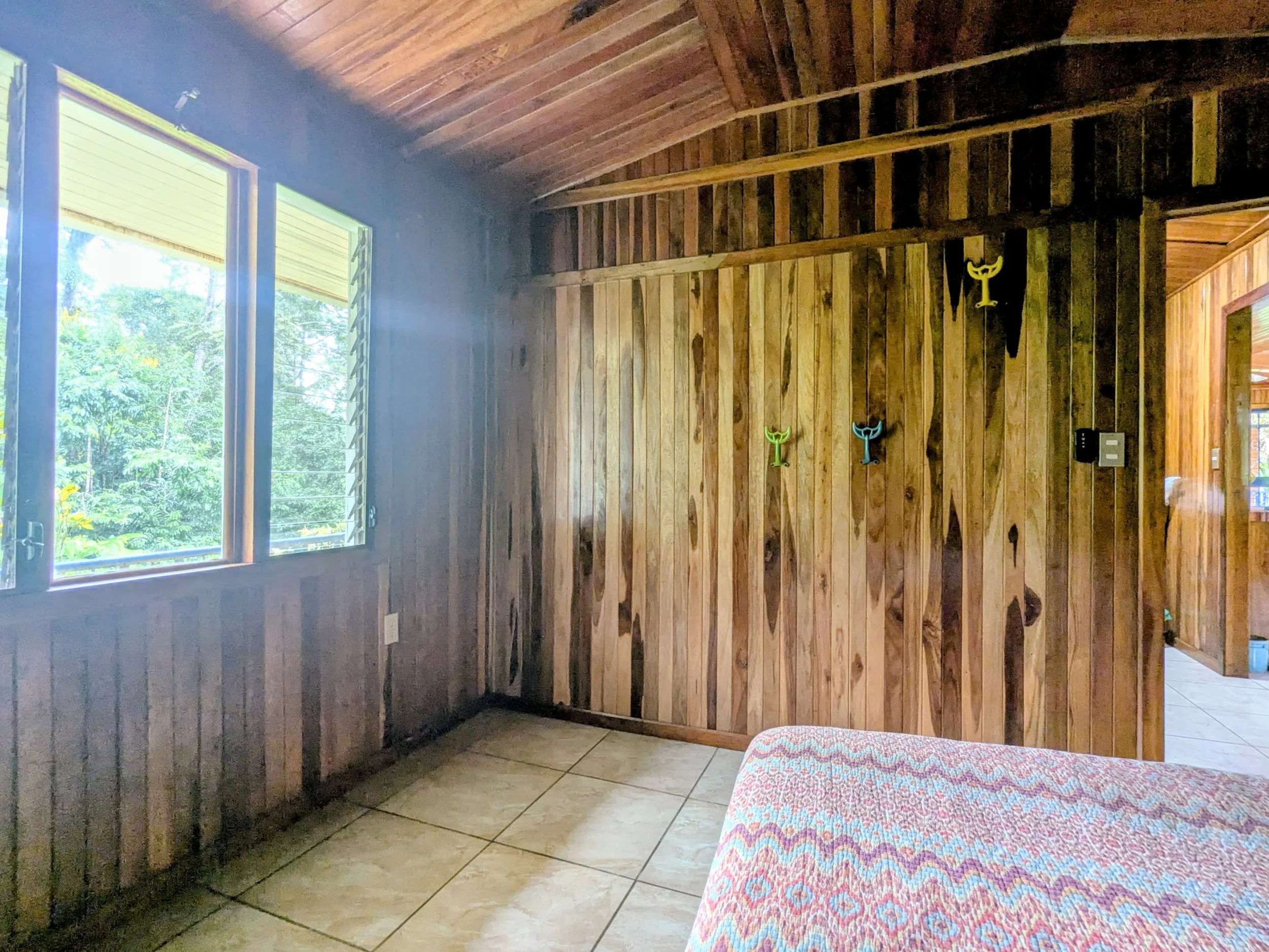A room with wooden paneled walls and ceiling, a window on the left side showing greenery outside, and colorful hooks mounted on the wall. Part of a patterned couch is visible in the foreground.