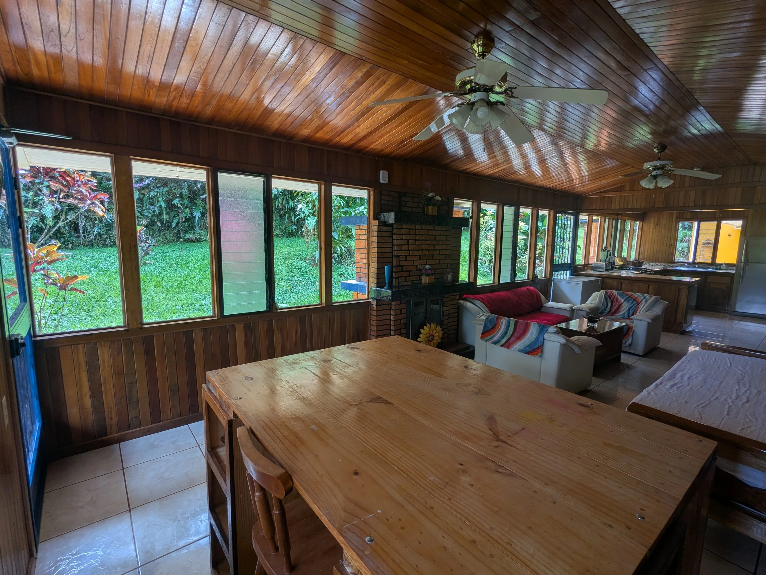 Living room with wooden walls and ceiling, large windows overlooking a garden, ceiling fans, and a brick fireplace. Furniture includes white sofas with colorful blankets, a wooden dining table and chairs, and a kitchen area in the background.