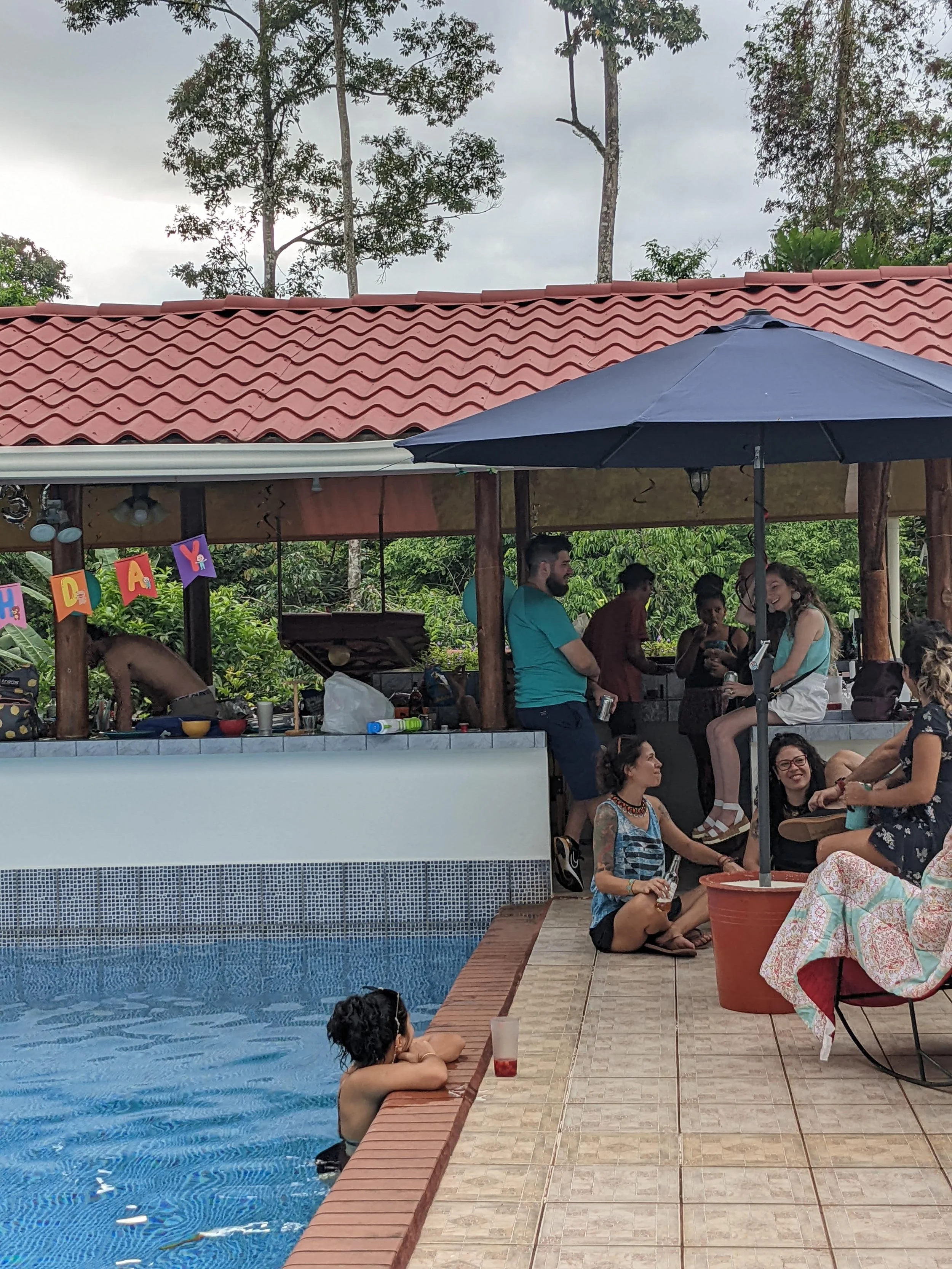 Group of people relaxing and socializing around a swimming pool with a covered patio, umbrella, and lush greenery in the background during daytime.
