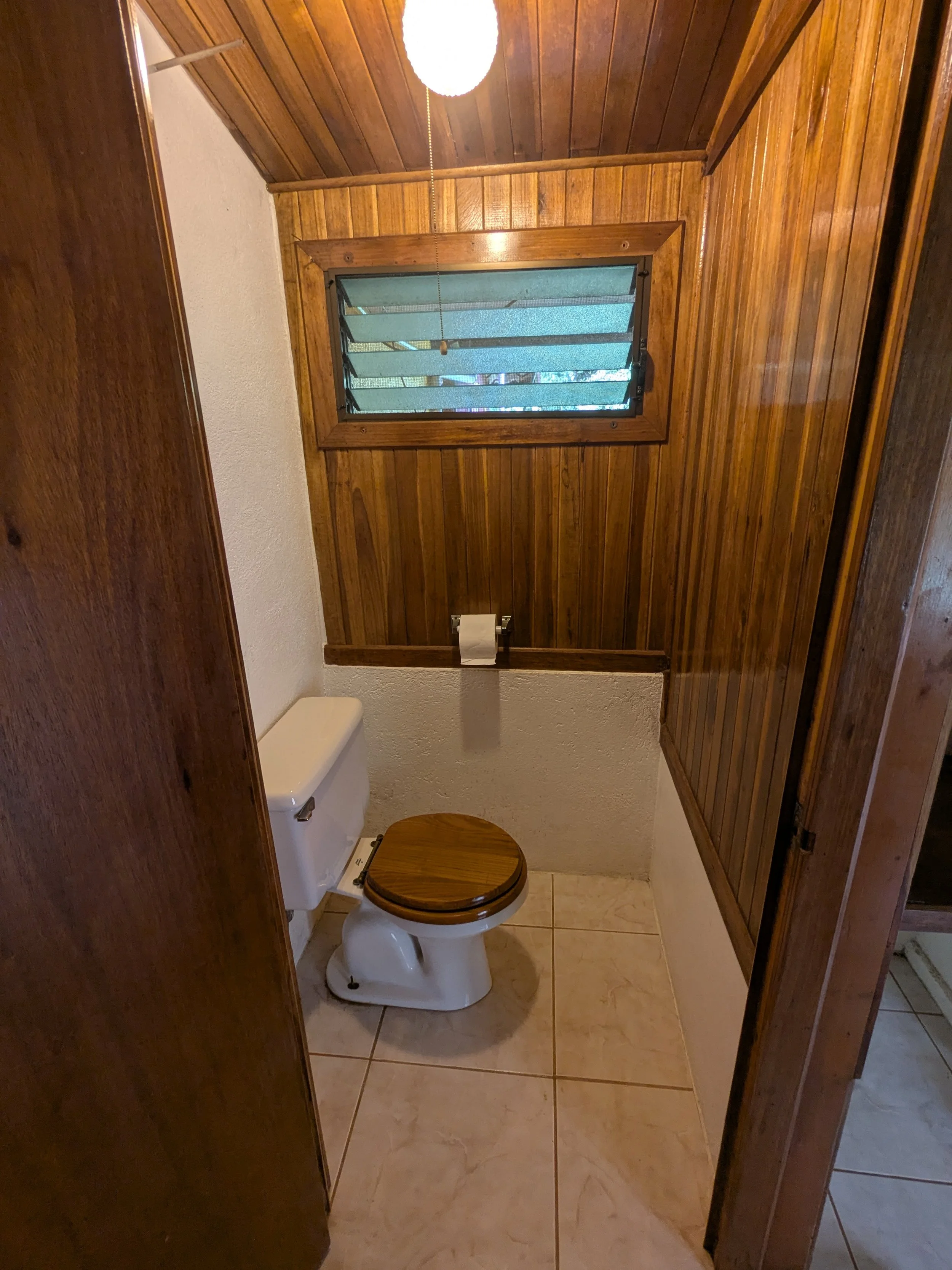 A small bathroom with wood-paneled walls and ceiling, a small window with horizontal louvers, and a white toilet with a wooden seat and lid.