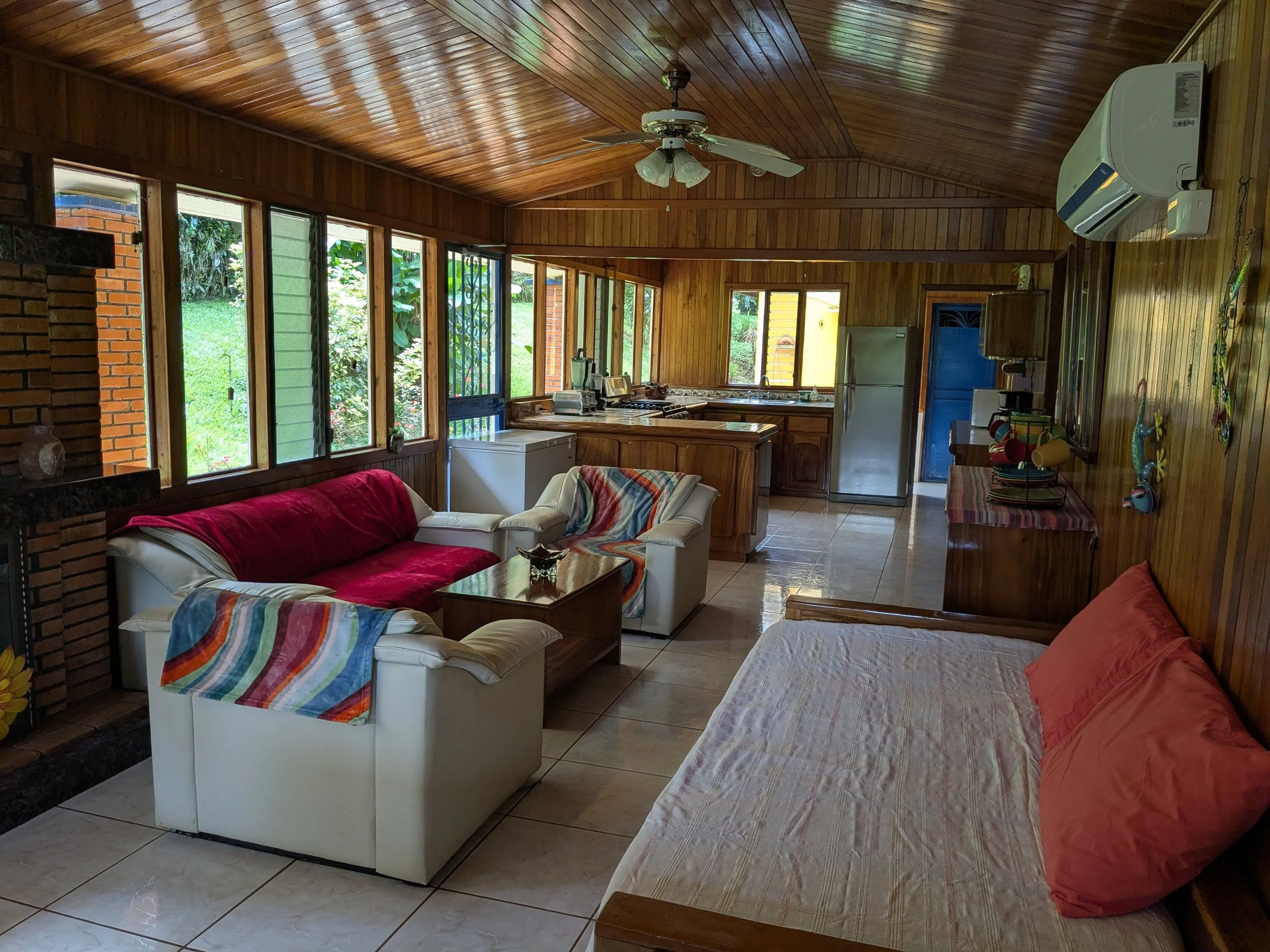 A cozy living room with wooden paneled walls and ceiling. It has a white sofa with a red cover and a matching armchair, a coffee table, and a daybed with a pink pillow. Large windows with outside greenery, a ceiling fan, air conditioner, and a kitche