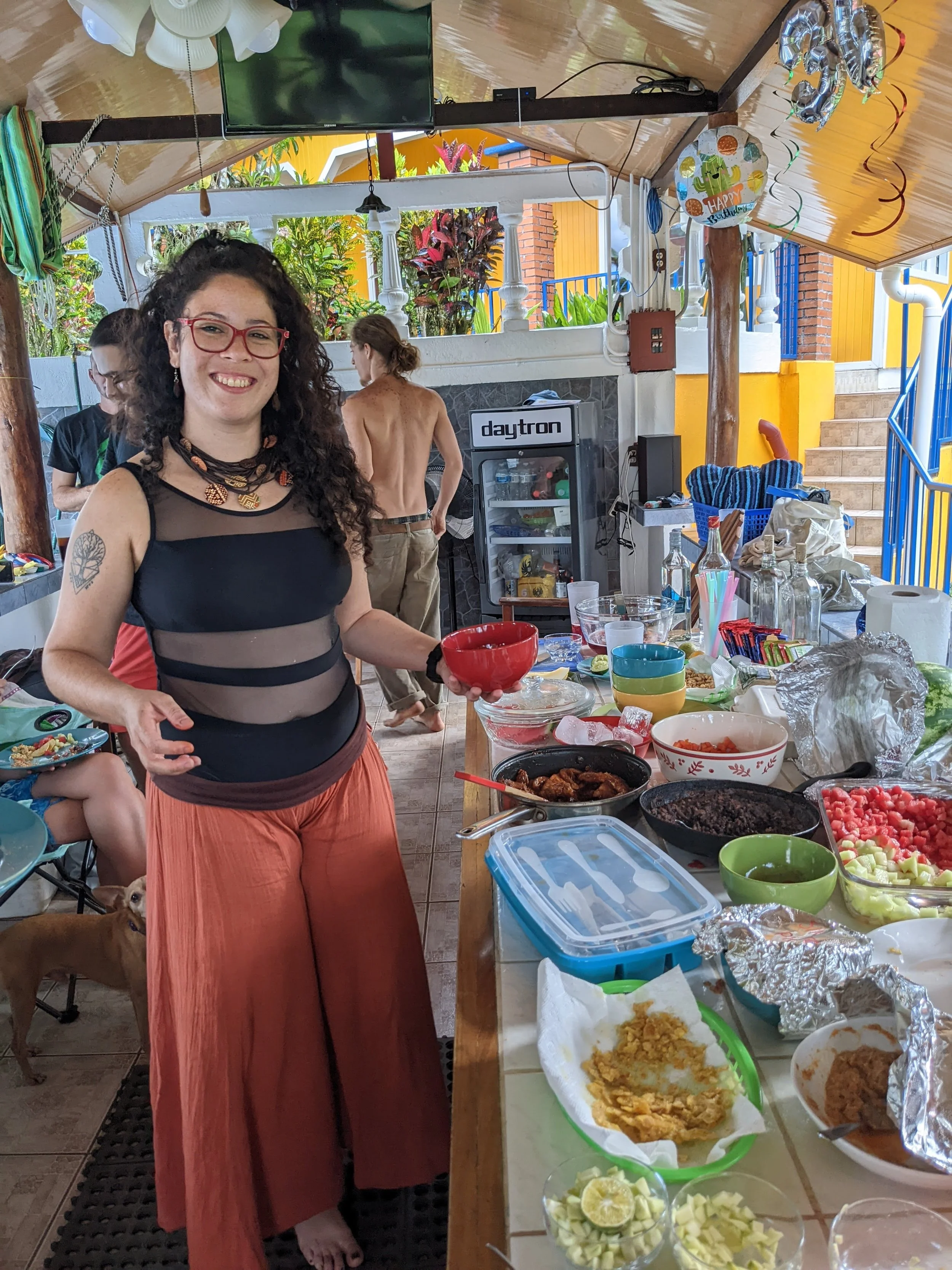 A woman with curly hair and red glasses smiling at a barbecue or outdoor party, holding a red bowl, with food and drinks on the table in front of her, and other people and a dog in the background.