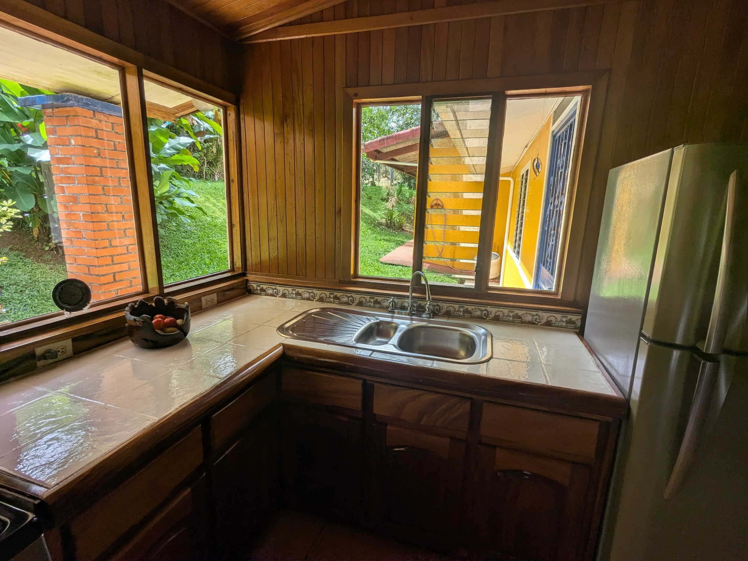 Kitchen with wooden cabinets, tiled counter, stainless steel double sink, and large windows showing greenery outside.