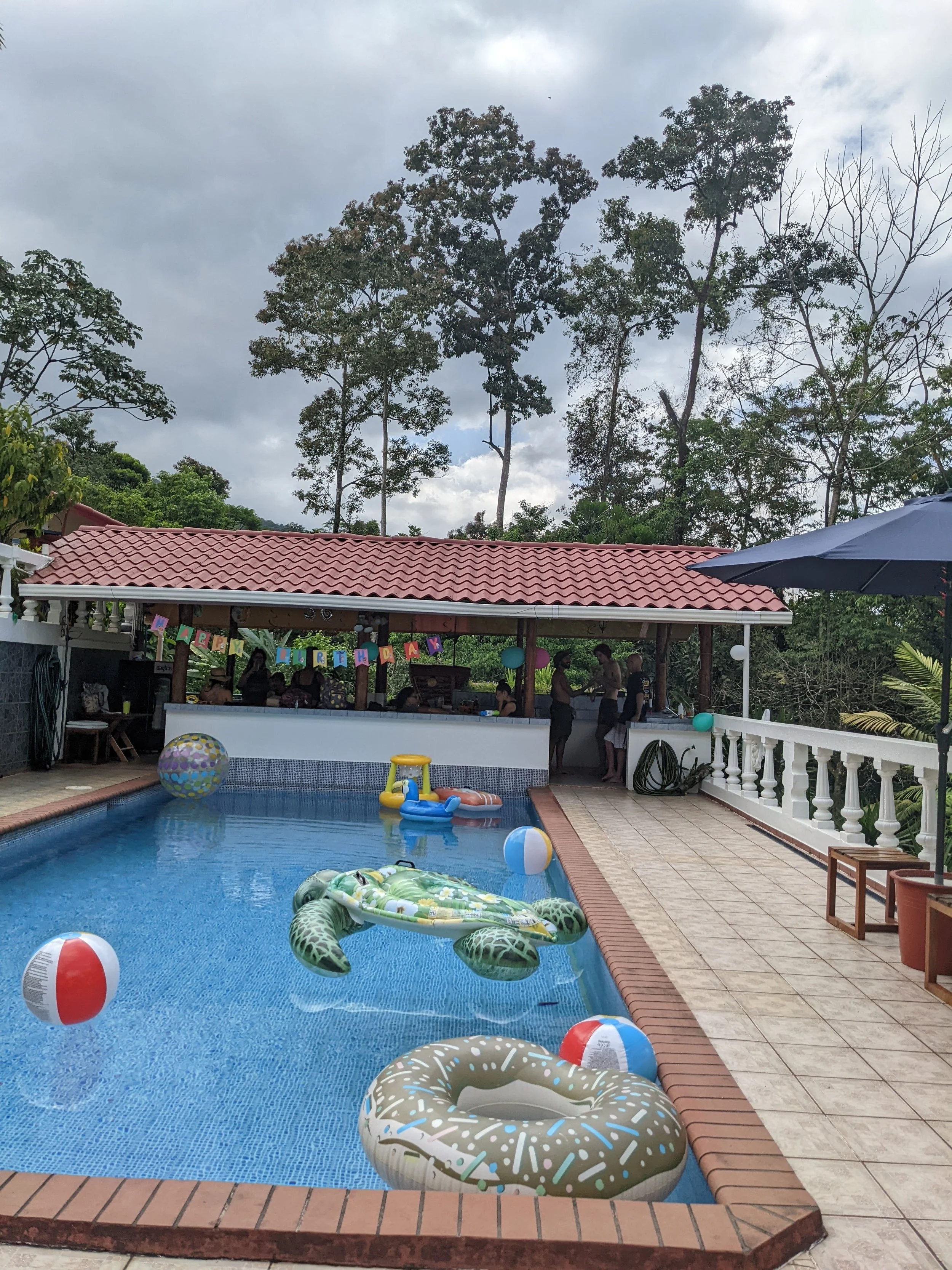 A pool party scene with a swimming pool filled with inflatable floaties, including a turtle and donut, and beach balls. There is a covered patio area with people, and a string of colorful banners hanging across it that reads 'Happy Birthday.' The bac