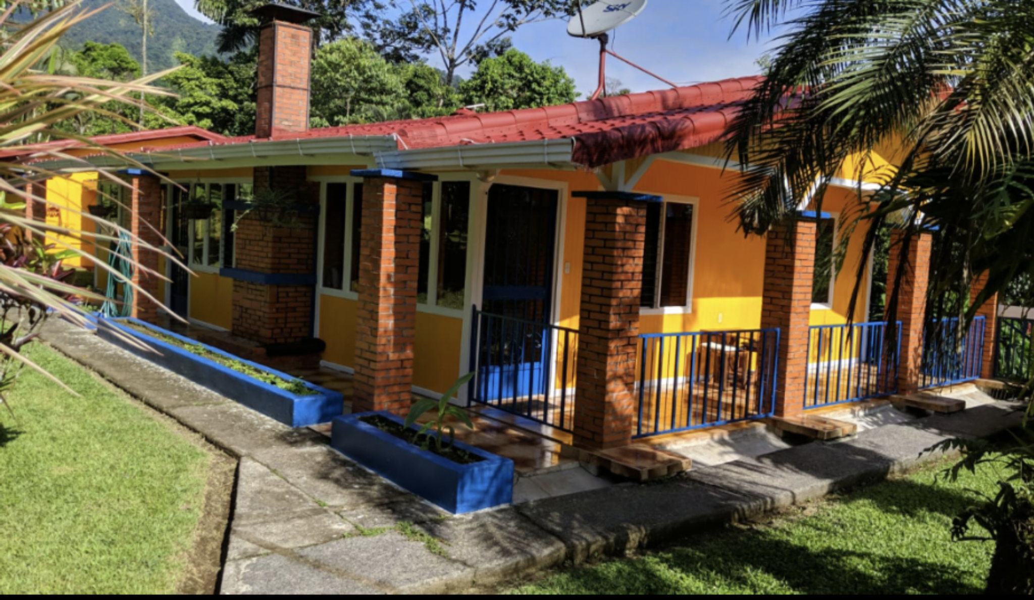 Colorful house with yellow walls, red roof, brick pillars, and blue railings, surrounded by greenery and plants. Costa Rica home and airbnb for sale in the jungle.