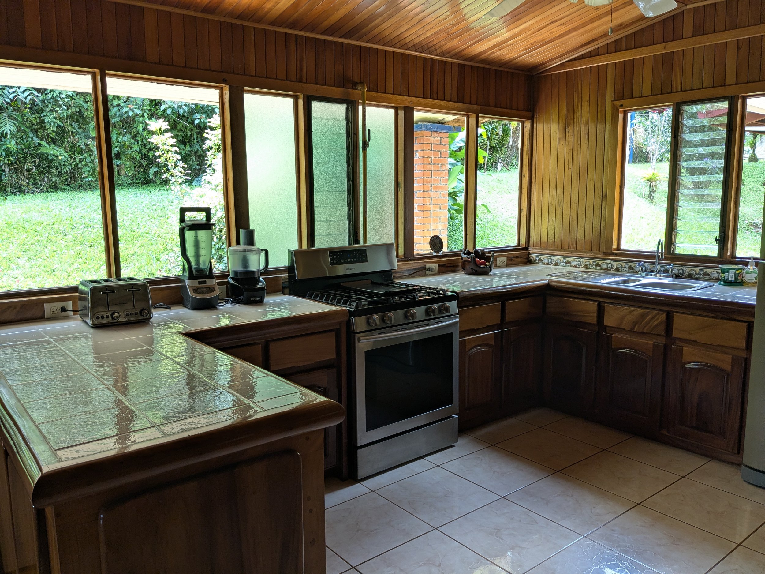 Kitchen with wooden cabinets, tiled countertops, and large windows overlooking a green yard with trees and grass.