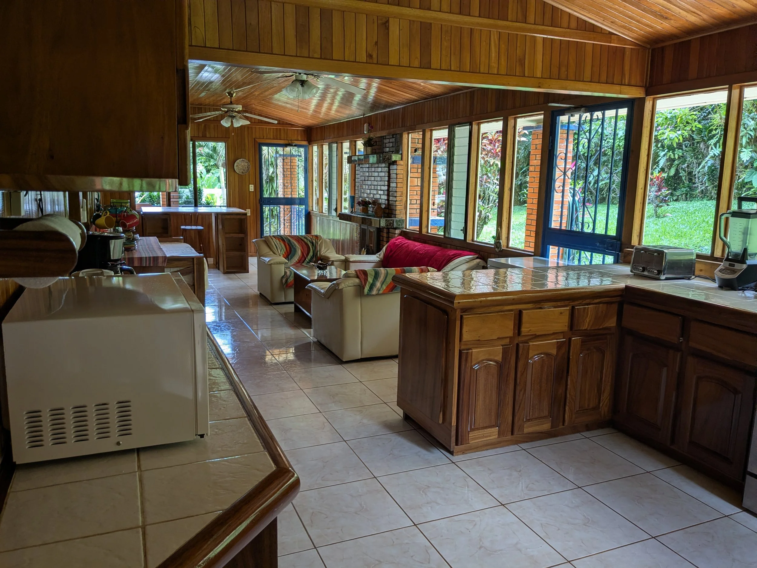 Kitchen and living room with wooden walls and ceiling, tiled floor, white sofas with colorful blankets, windows with greenery outside, and a fireplace.