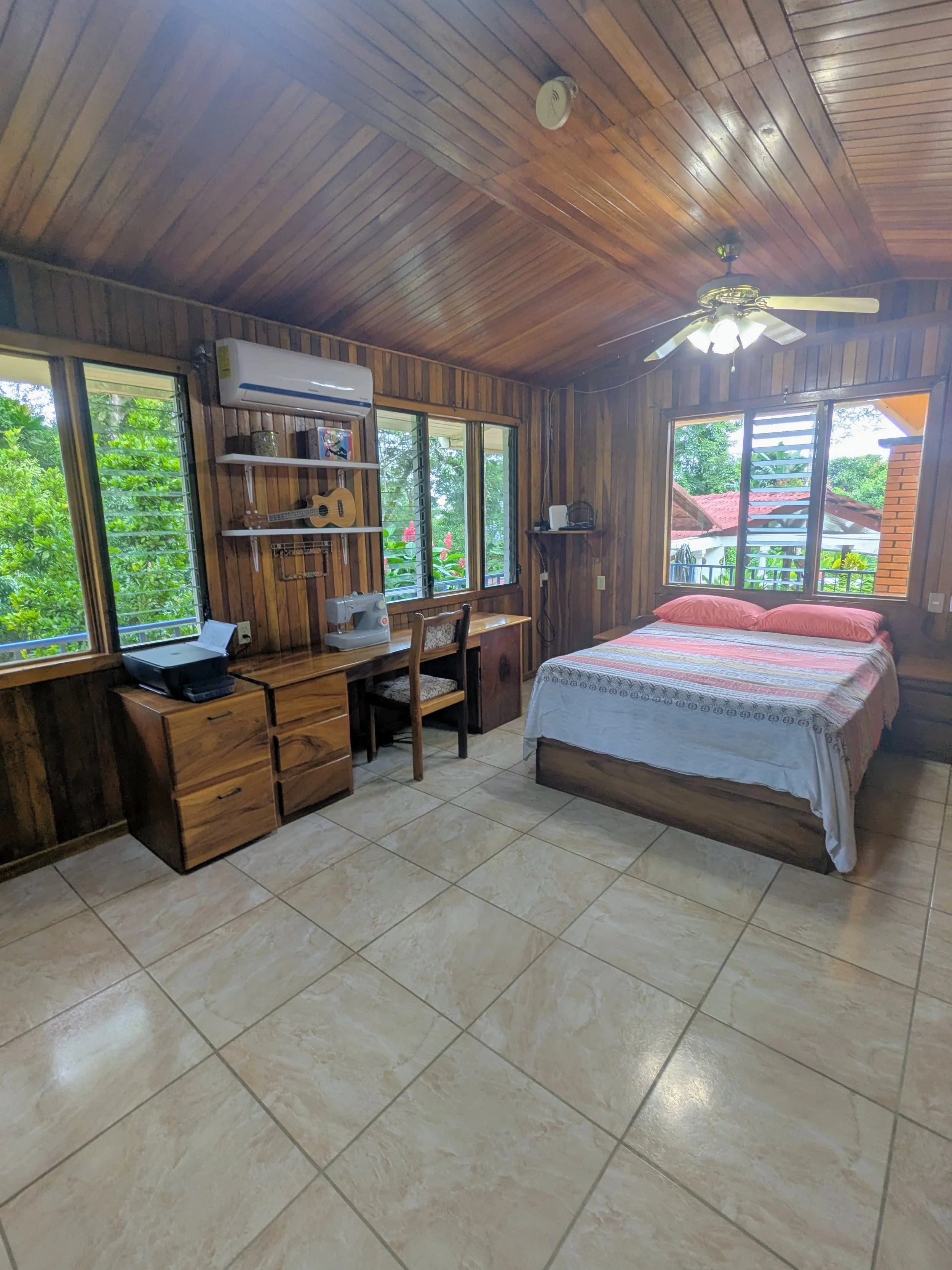 A bedroom with wooden walls and ceiling, a bed with a colorful bedspread and pink pillows next to a window, a desk with a sewing machine and printer, and shelves with a guitar and framed picture, illuminated by natural light. Costa Rica home and airb