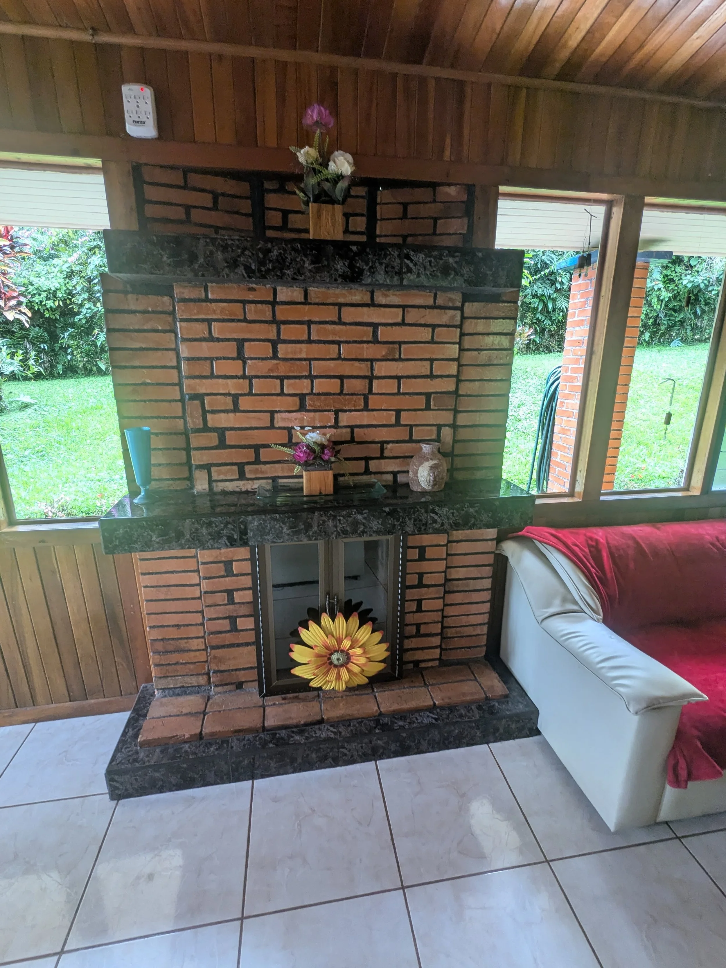 Indoor brick fireplace with black marble mantel decorated with flower arrangements and a decorative ceramic jar, flanked by large windows with views of a green garden, next to a white sofa with a red blanket.