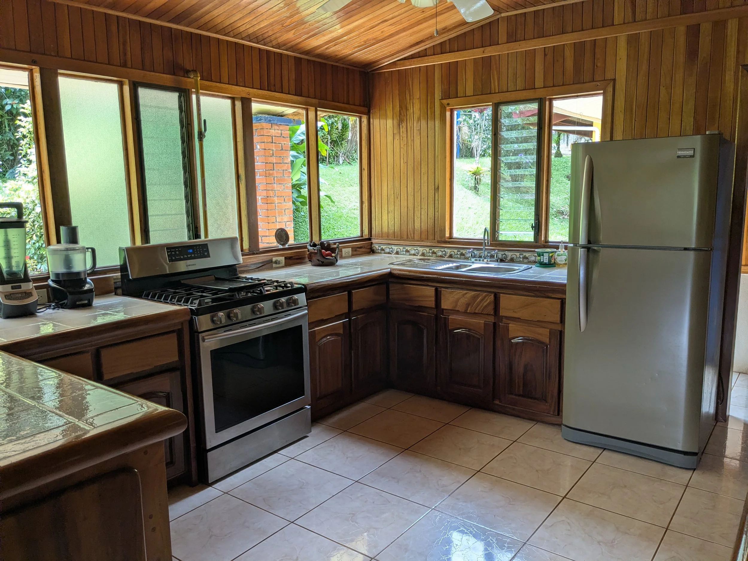 Kitchen with wooden cabinets, tiled counters, stainless steel stove, refrigerator, blender, and sink with windows showing a lush green outdoor area.