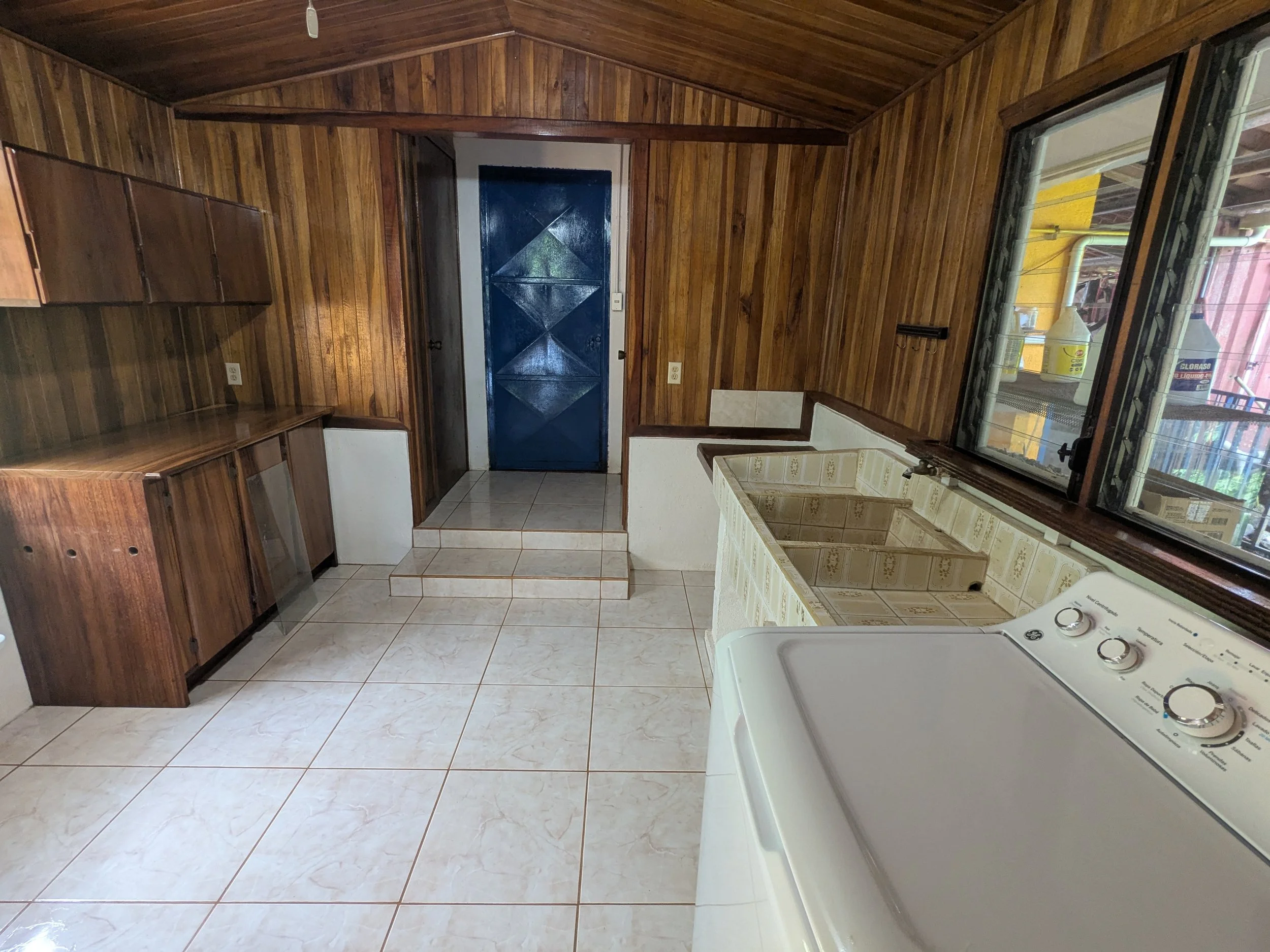 Laundry room with wood-paneled walls, white tile floor, a white top-load washing machine, a tiled sink with beige and brown patterns, a window with a view of a yellow building, and a blue door at the back.