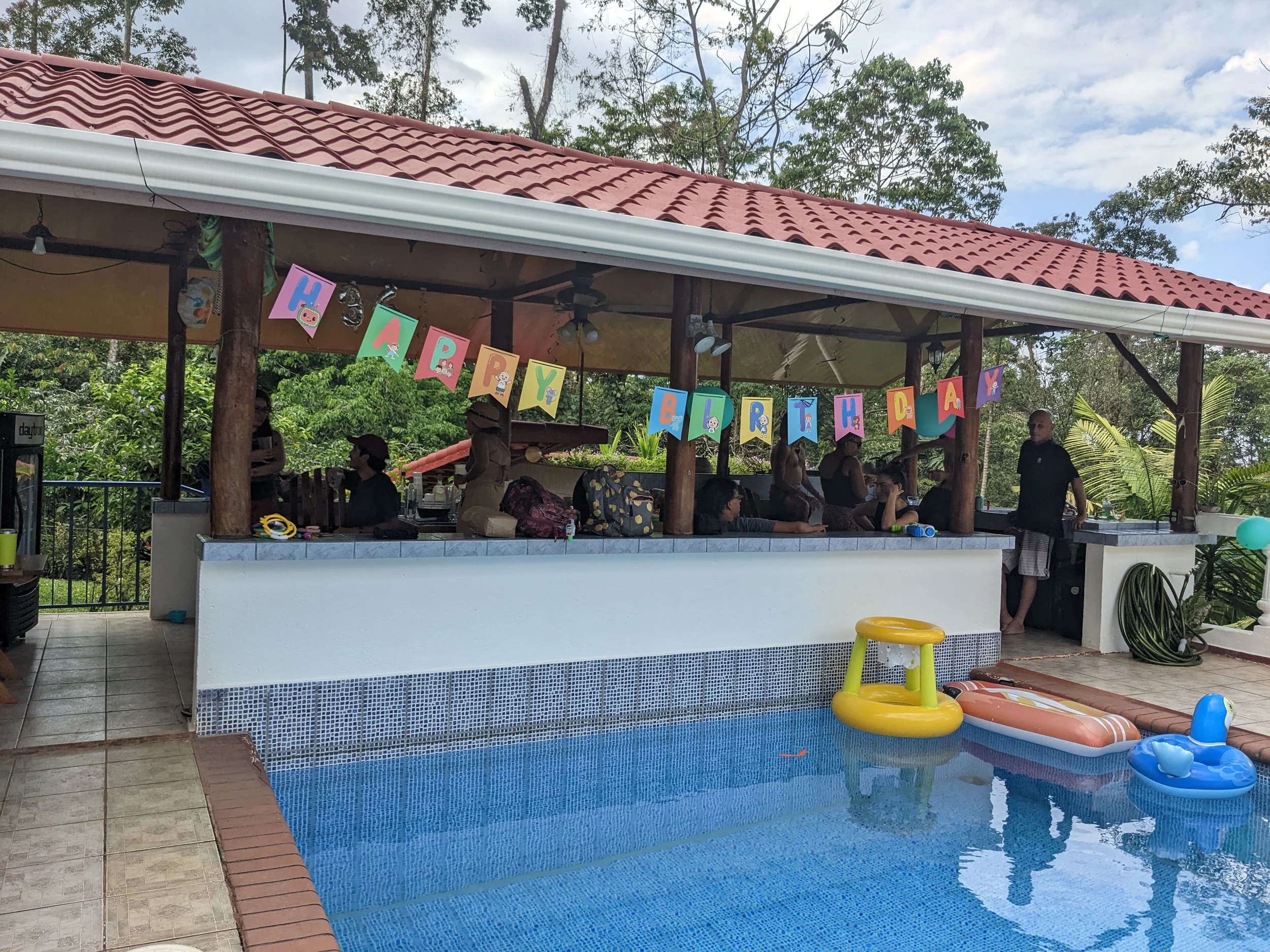 Poolside area decorated for a birthday celebration with a colorful "Happy Birthday" banner, inflatable pool toys, and a partially visible bar or serving area with people.
