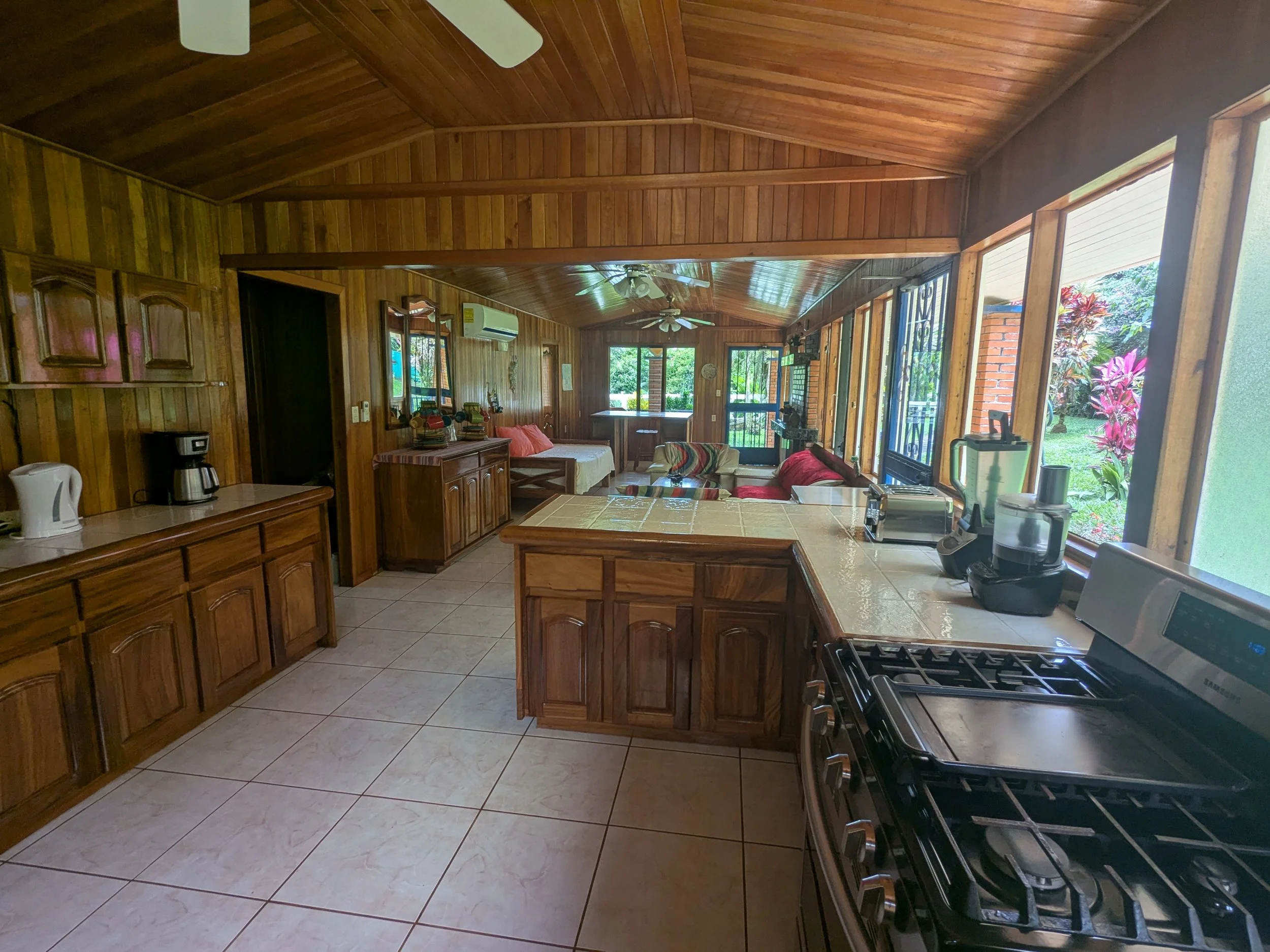 Interior view of a wooden kitchen and living area with tile flooring, a stove, coffee maker, and various kitchen appliances, with large windows revealing a garden outside.