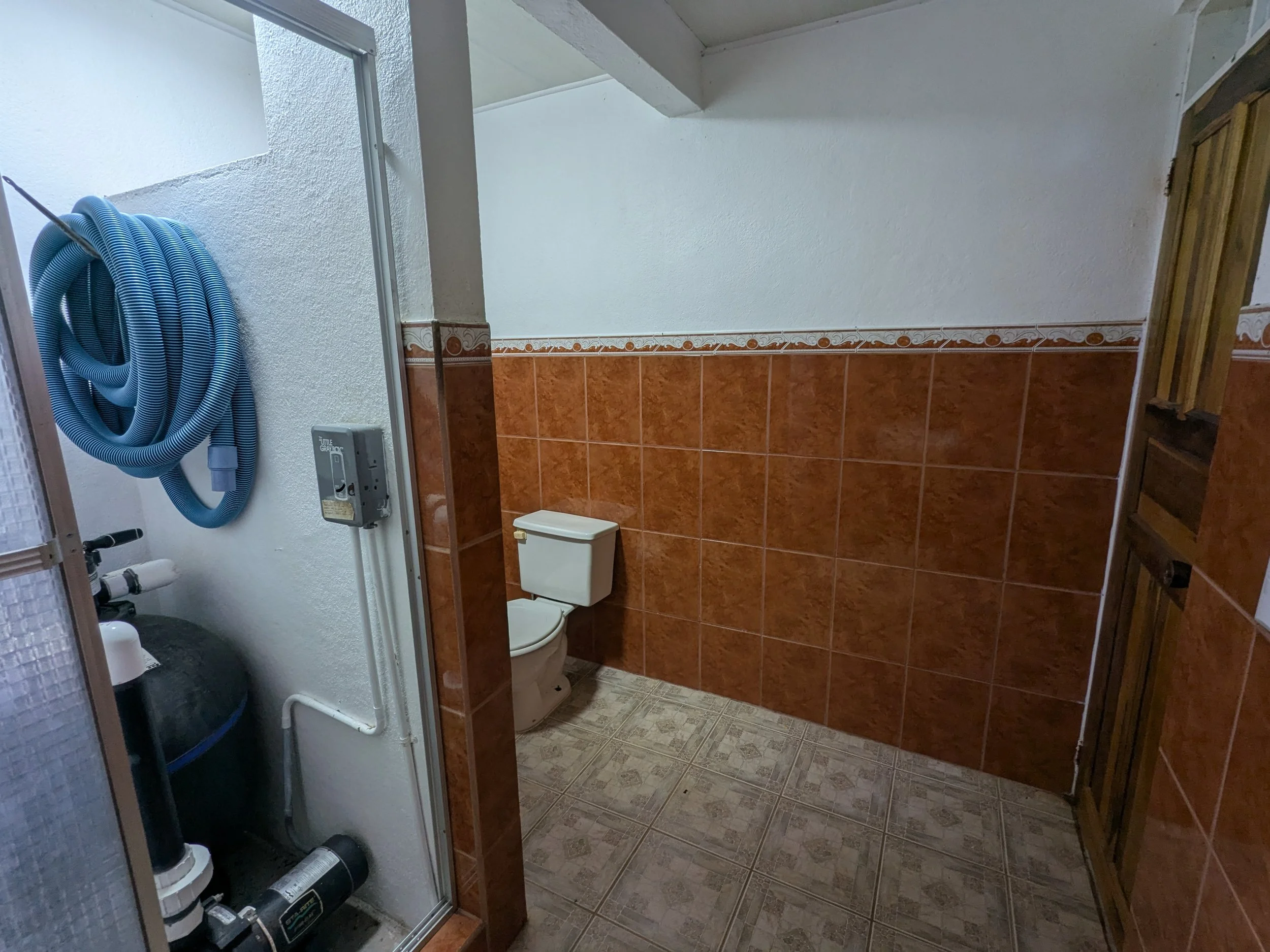 Bathroom with a toilet, brown tiles on the lower half of the wall, white upper wall, a wooden door, and a water hose stored on the left side.