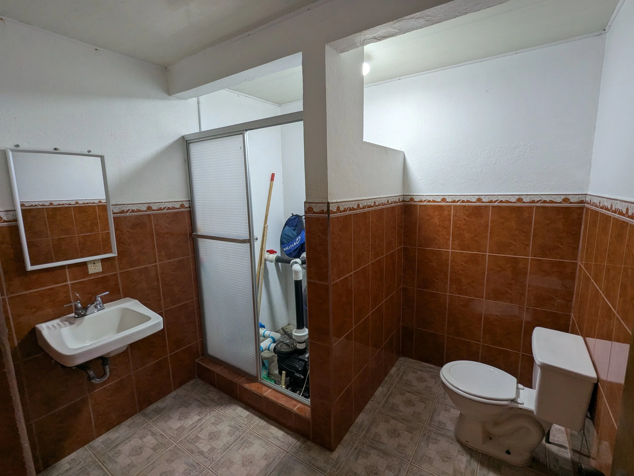 A bathroom with a small sink, a toilet, and a divided shower area. The walls are tiled in brown with decorative border tiles, and the floor has patterned tiles. There's also exposed plumbing and cleaning supplies behind the shower.