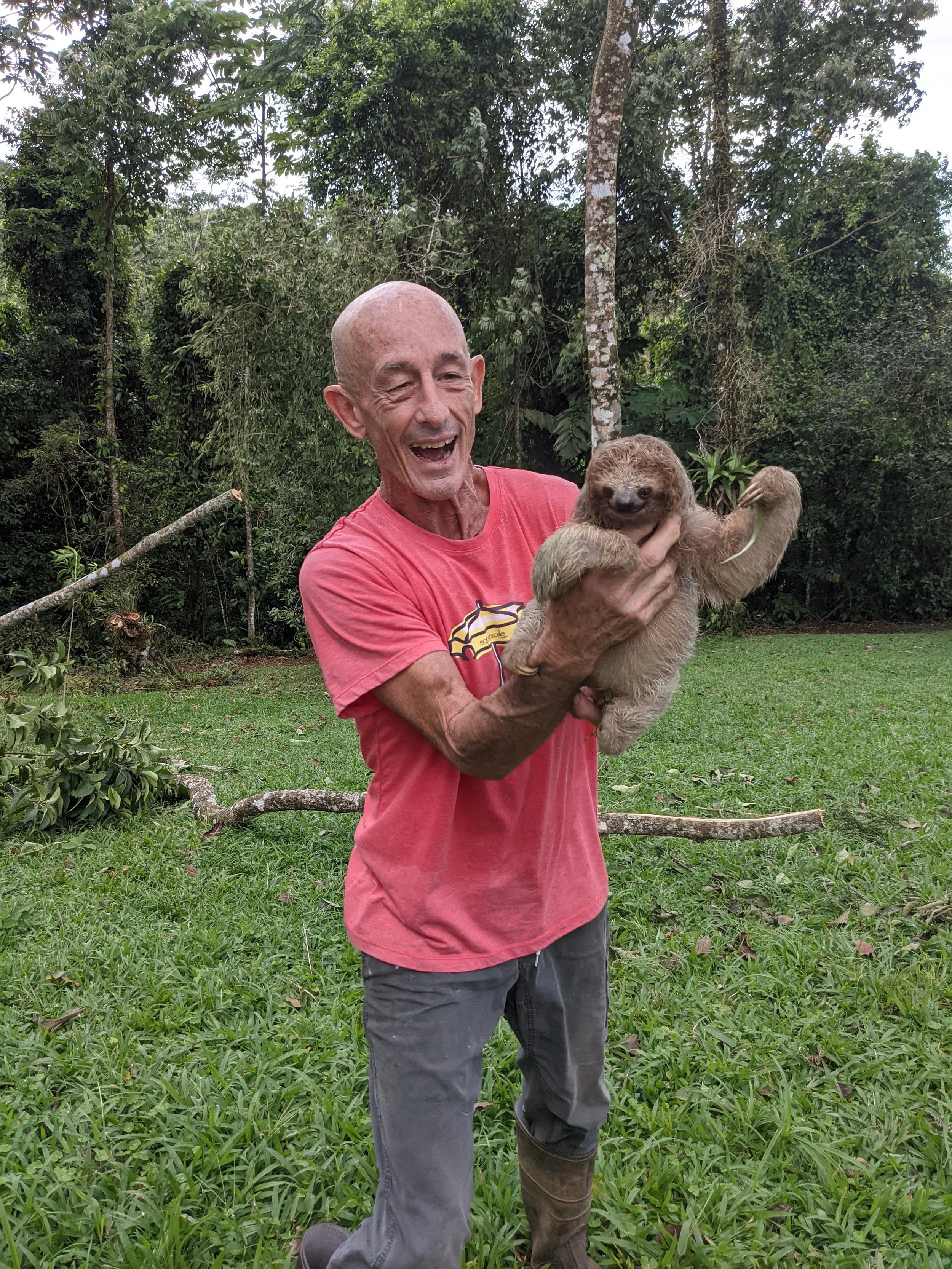 An older man in a red t-shirt and dark pants holds a baby sloth in a lush, green outdoor setting, smiling.