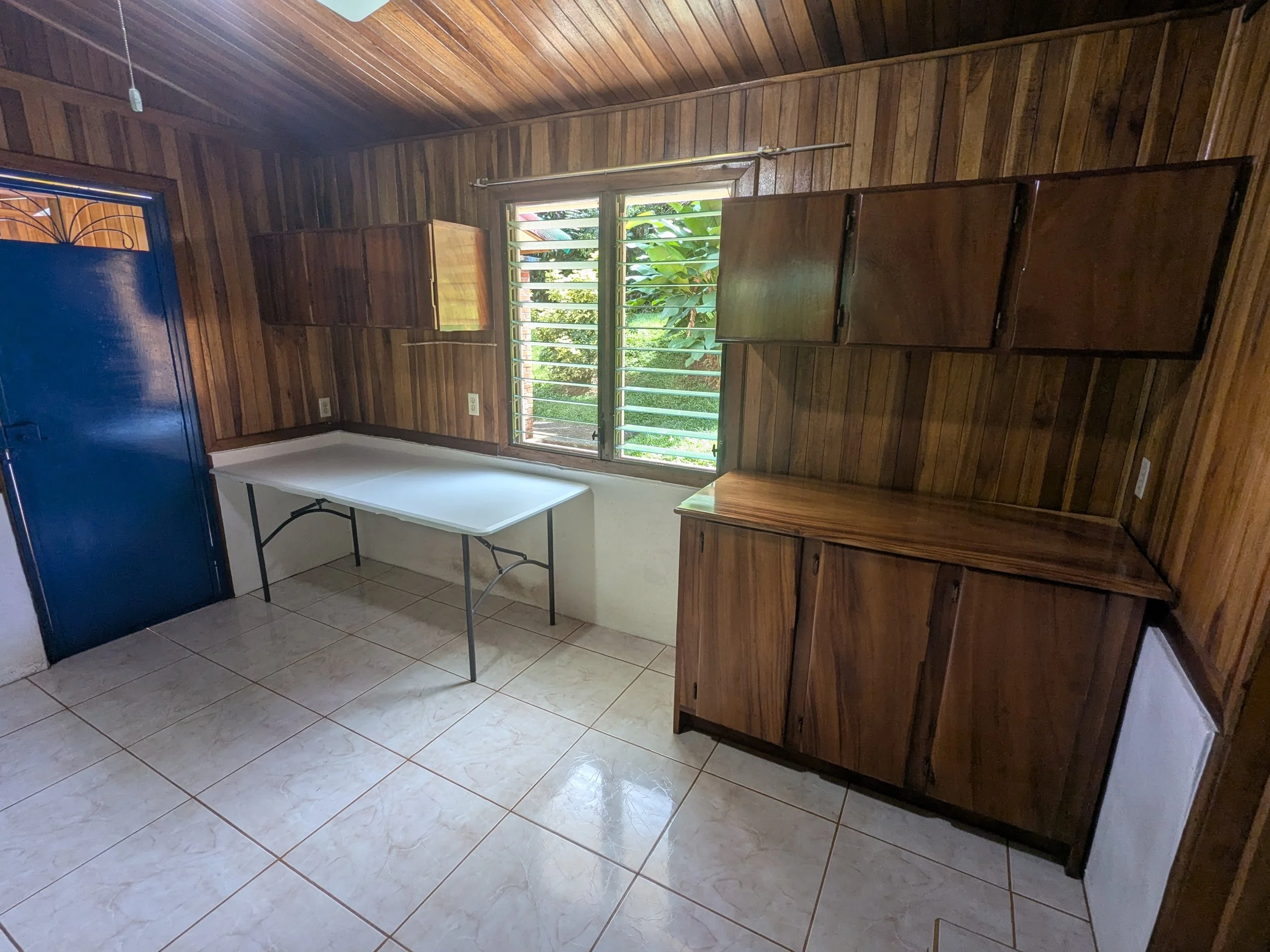 A small room with wooden panel walls and a tiled floor. There is a window with horizontal metal bars showing green foliage outside. The room has wooden cabinets and a white foldable table, with a dark blue door on the left side.