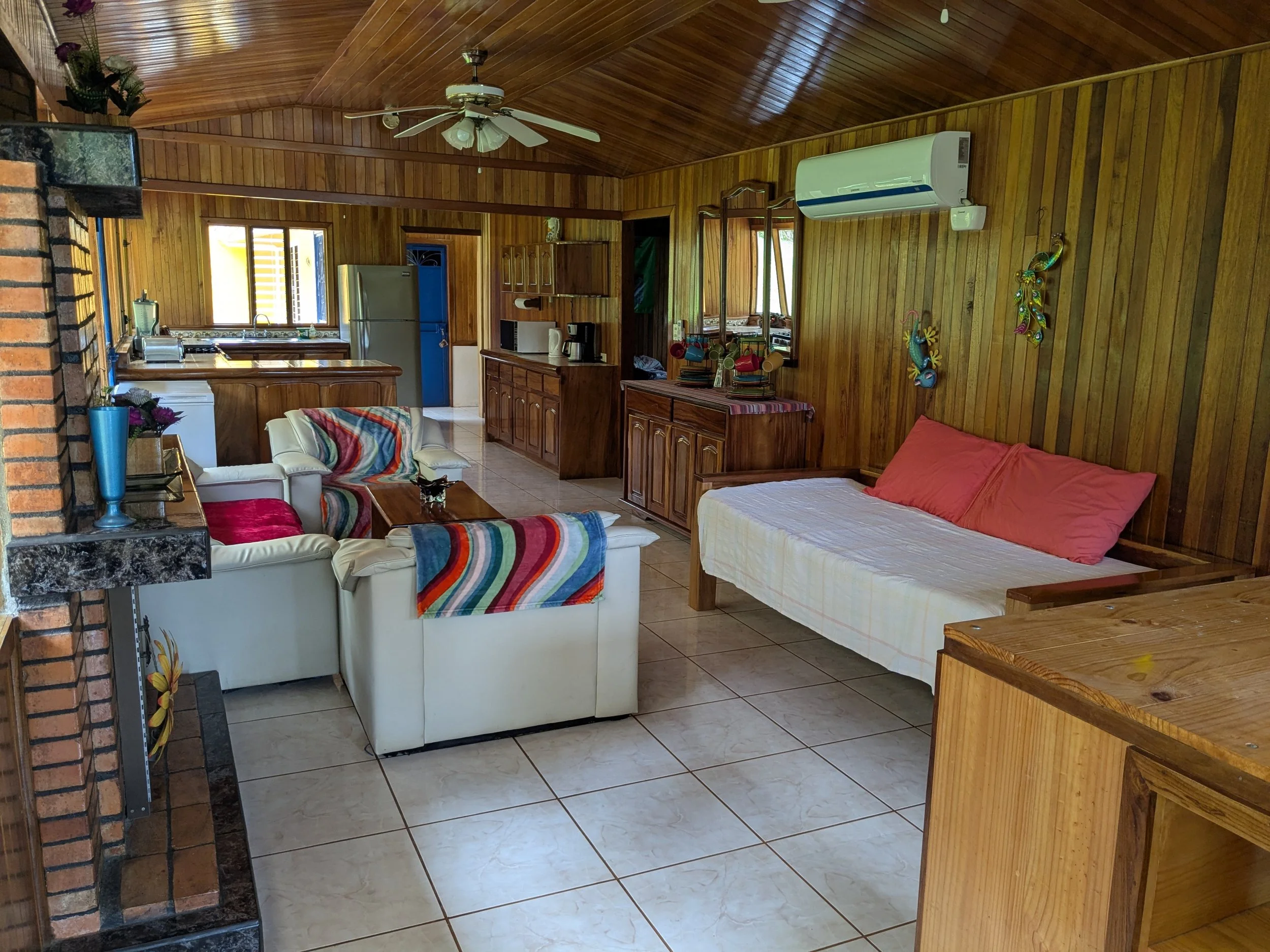Living room with heart-shaped bed, white sofa with colorful blankets, kitchen area with refrigerator, stove, and microwave, and wooden wall paneling.