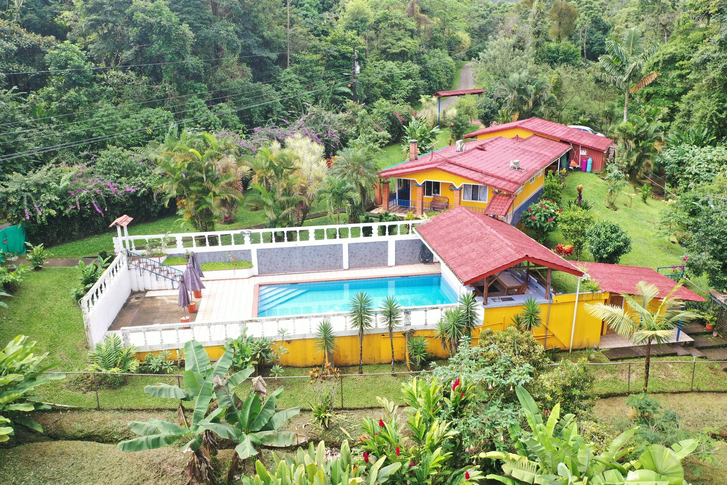 Aerial view of Costa Rica home in the jungle. Colorful house with red roof, surrounded by lush greenery, trees, and a backyard with a swimming pool, umbrellas, and outdoor furniture.