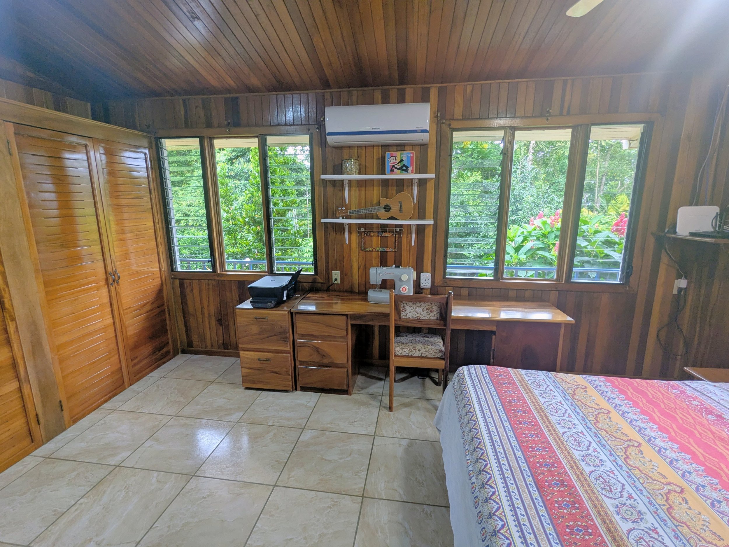 A cozy wooden bedroom with a bed, window view of greenery, a work desk with a sewing machine, chair, and shelves with decorative items and a guitar. Costa Rica home and airbnb for sale in the jungle. Income earning property in Costa Rica