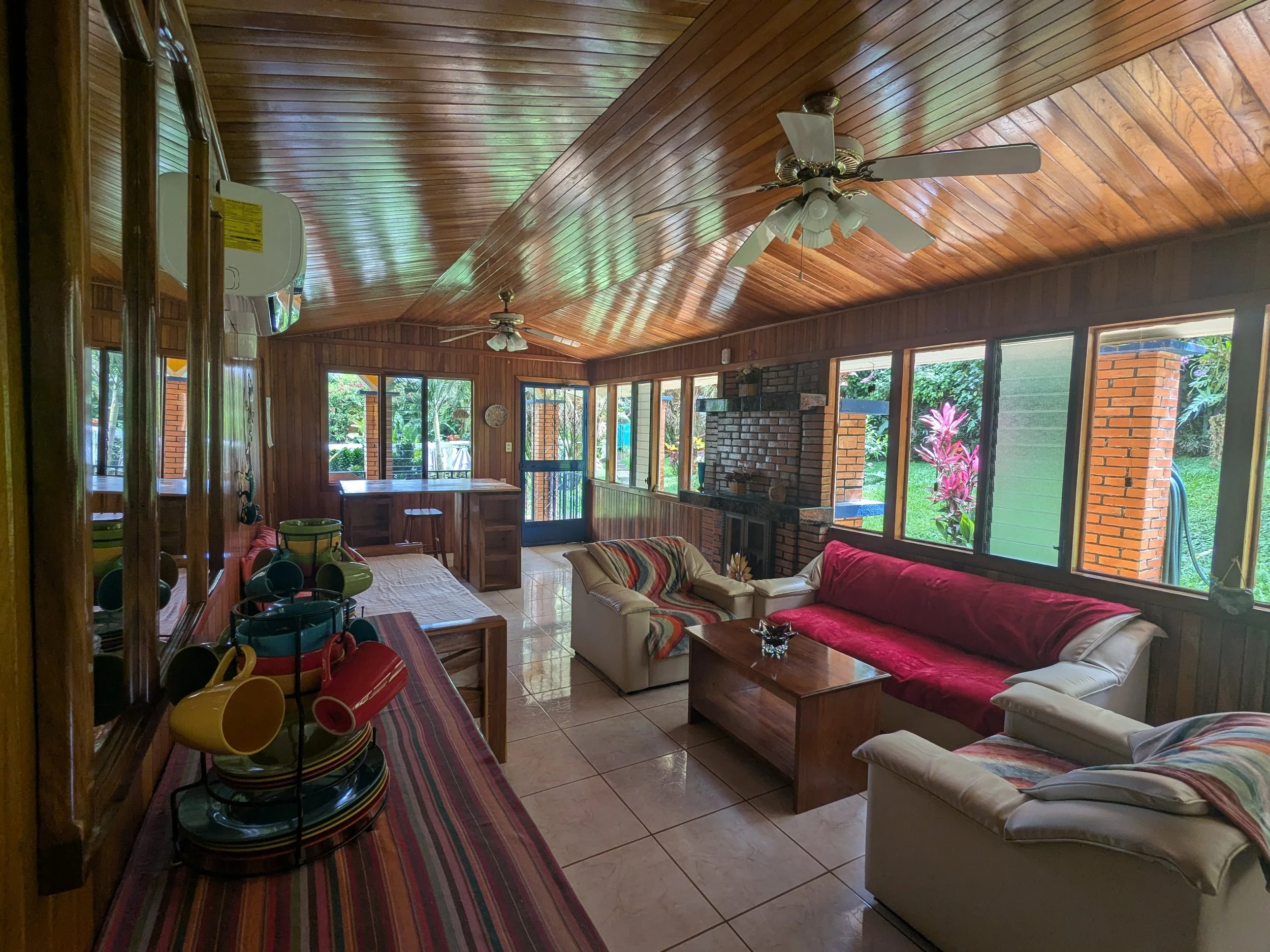 Interior of a sunroom with wooden walls and ceiling, ceiling fans, large windows showing greenery outside, red and beige sofas, a brick fireplace, and a wooden coffee table.