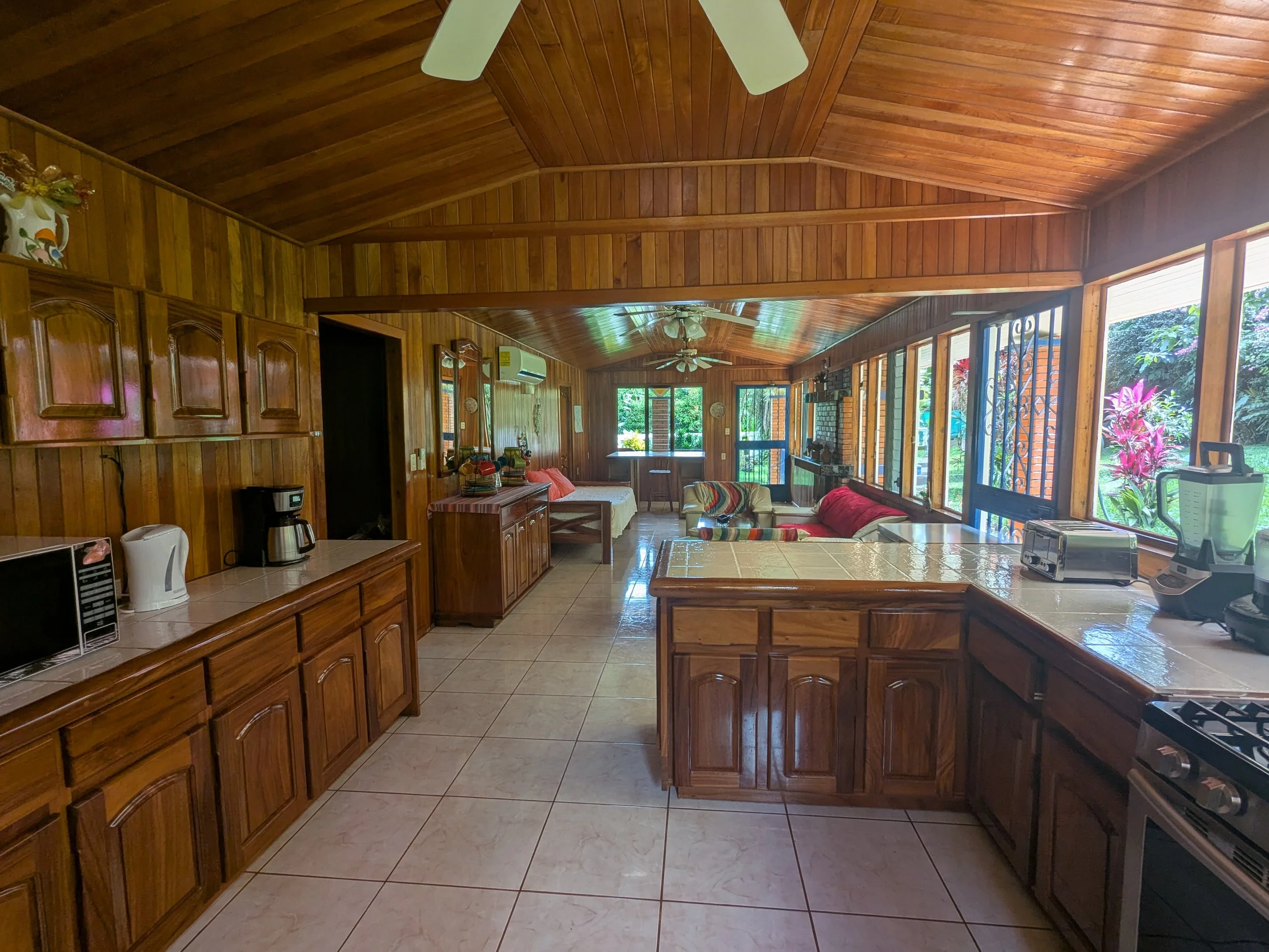 A spacious kitchen with wooden cabinets, tile countertops, and large windows overlooking a lush garden. A small appliance and coffee maker are on the counter. The open living area with sofas and ceiling fans is visible in the background.