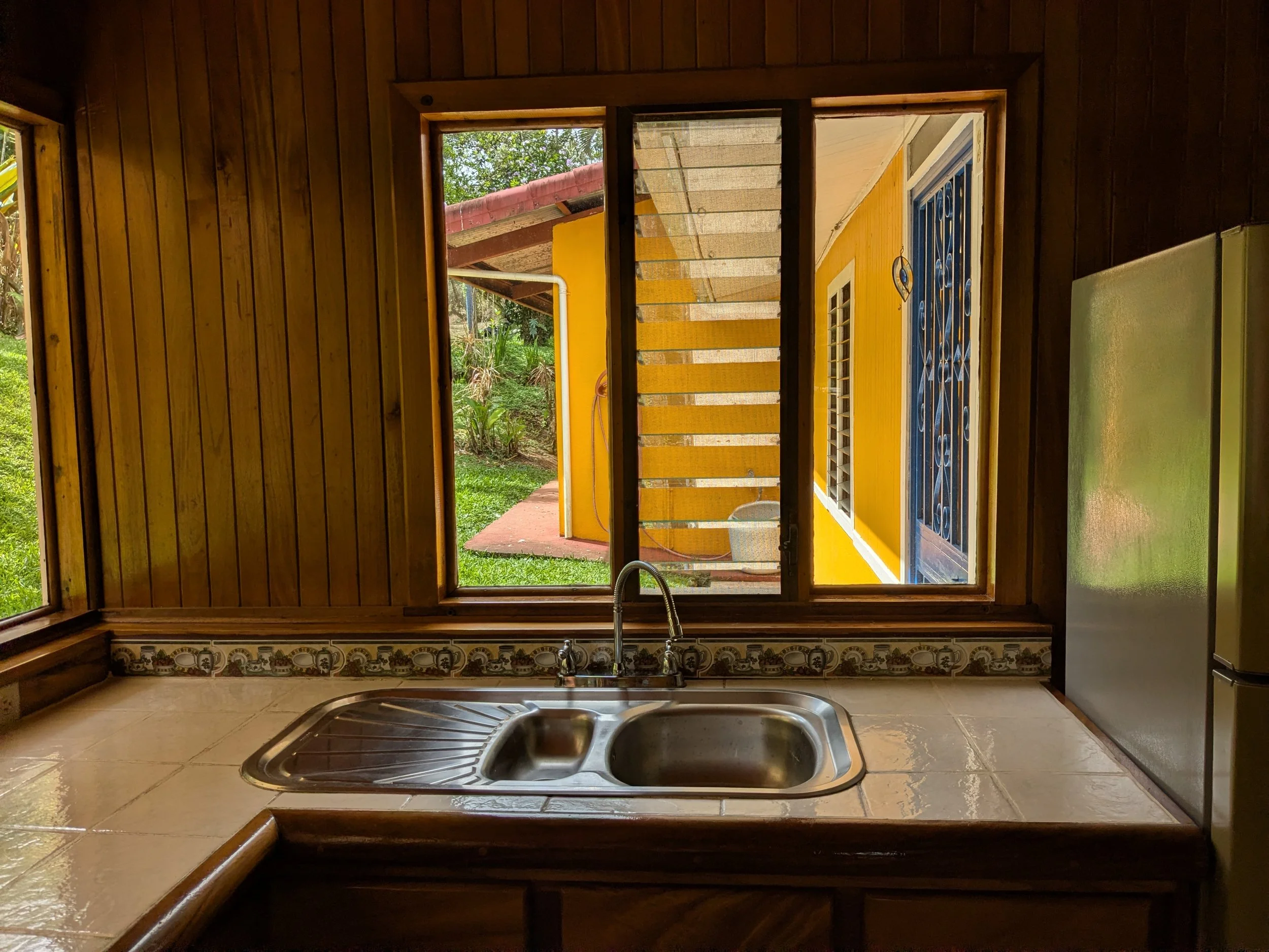 Kitchen with a double sink and a window showing a yellow exterior wall and greenery outside.