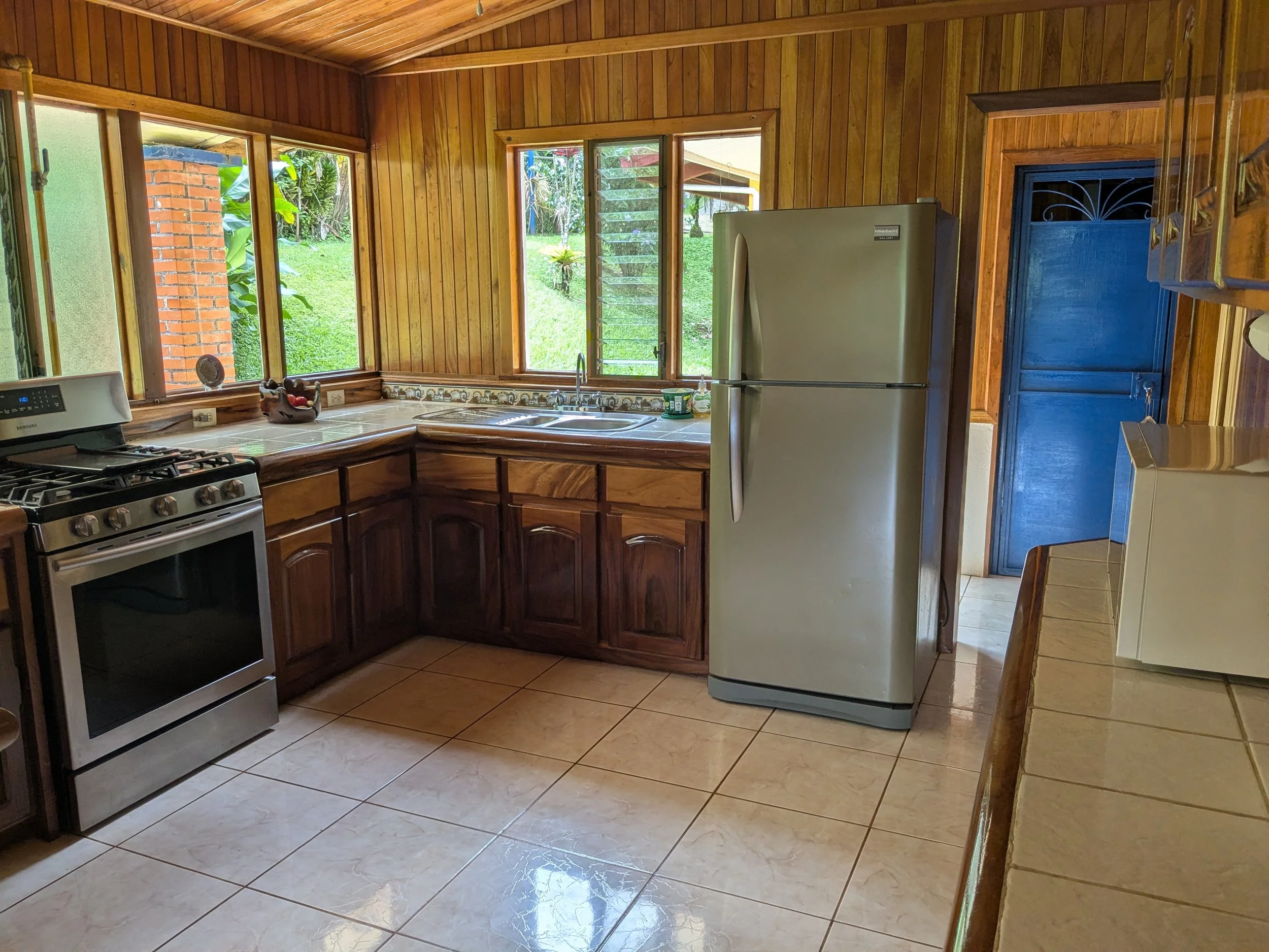 Kitchen with wooden walls, cabinets, and ceiling, including a windowed corner, stainless steel refrigerator, stove, and tiled countertops and floor.