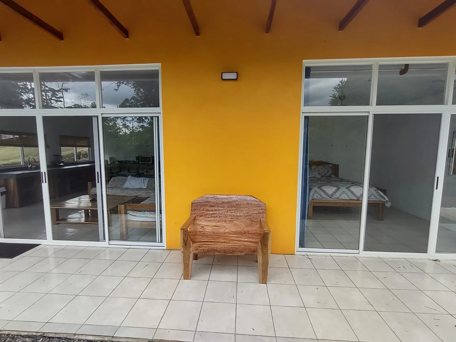 A building with yellow exterior walls, large glass sliding doors leading to bedrooms and a living area, and a single wooden chair on the tiled patio outside.