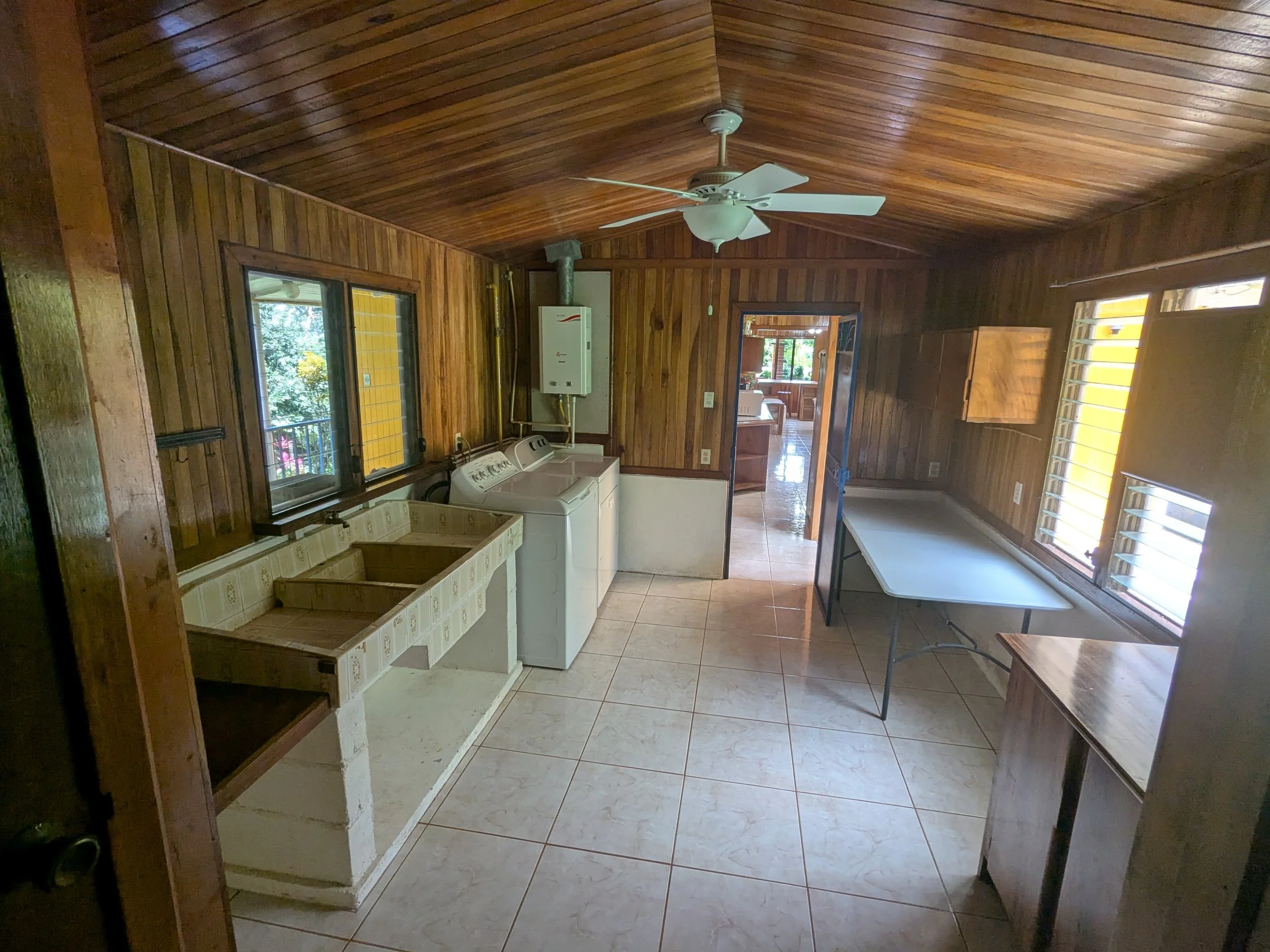 A laundry room with wood-paneled walls and ceiling, featuring windows and a ceiling fan. There is a utility sink with a tile countertop, a washing machine and dryer, a folding table, and a small wooden cabinet.