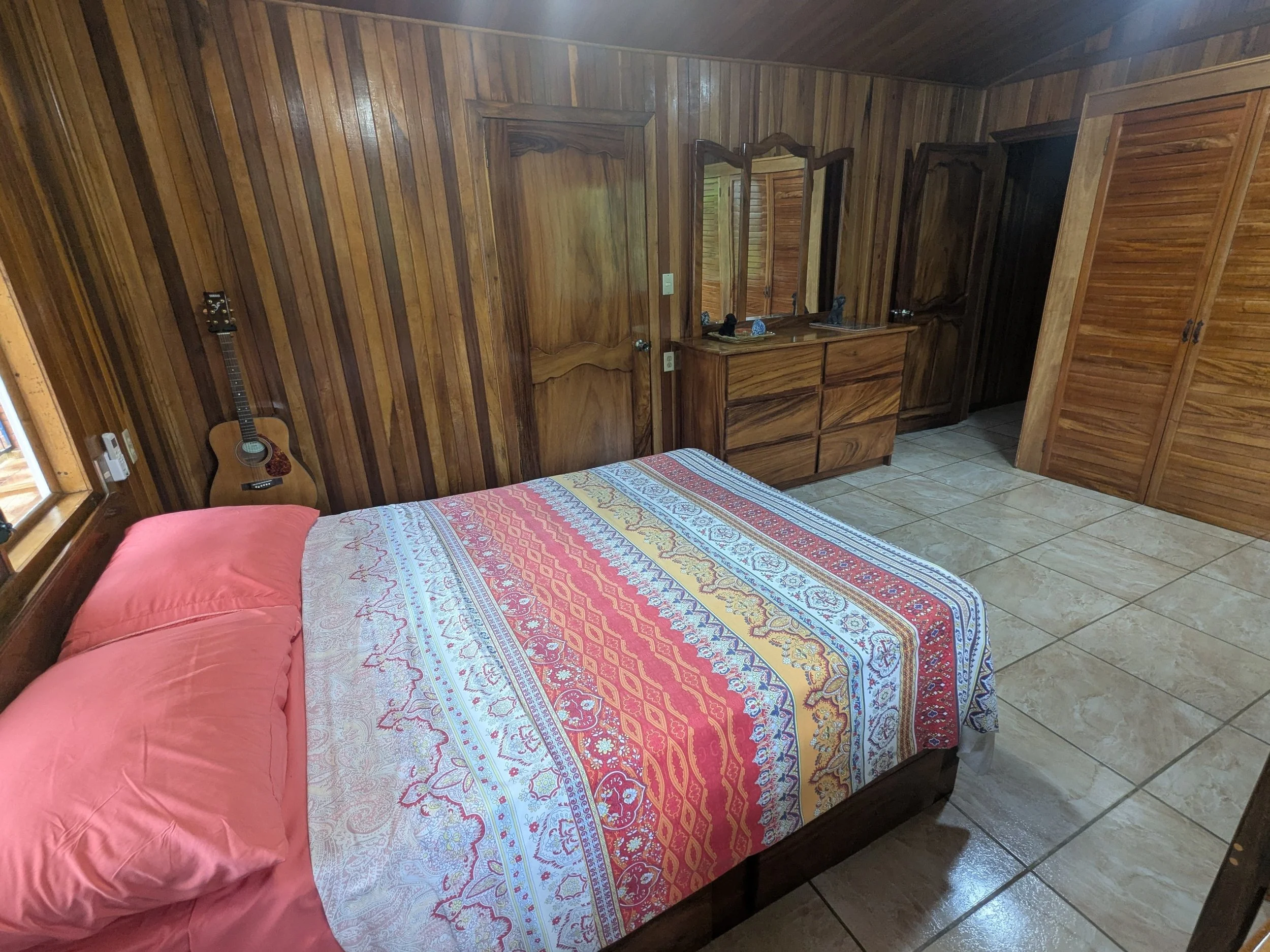 A bedroom with wooden paneled walls and beige tiled flooring. There is a double bed with pink pillows and a multicolored patterned bedspread. A small acoustic guitar is propped against the wall next to the bed. A wooden dresser and mirror are visible