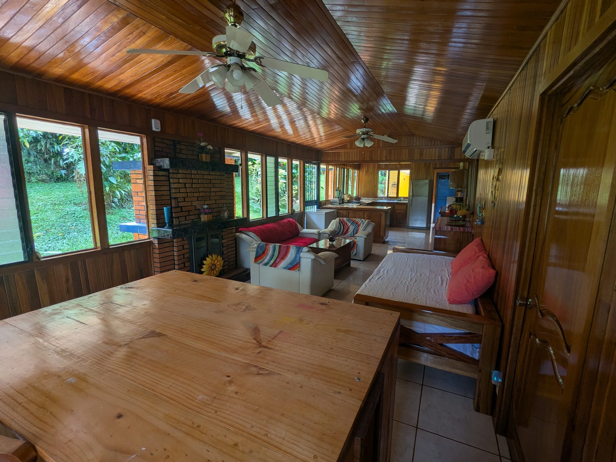 A cozy living room with wooden walls and ceiling, featuring large windows with a view of lush greenery outside. There is a brick fireplace, white sofas with colorful blankets, a dining table in the foreground, and a small kitchen area with a refriger