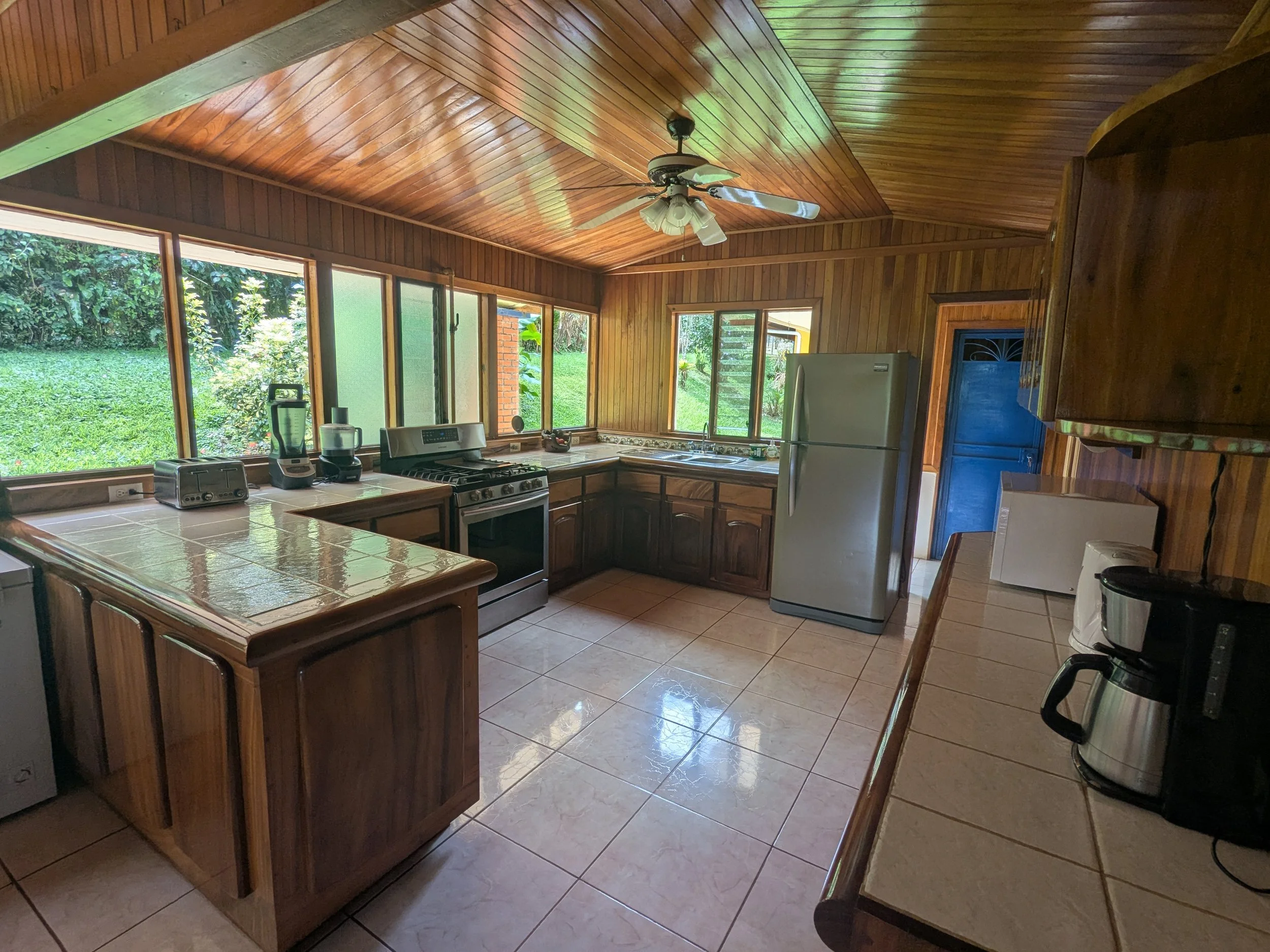 A wooden-paneled kitchen with large windows overlooking a grassy yard, featuring a white refrigerator, stove, microwave, coffee maker, blender, and toaster, with tiled countertops and ceiling fan.
