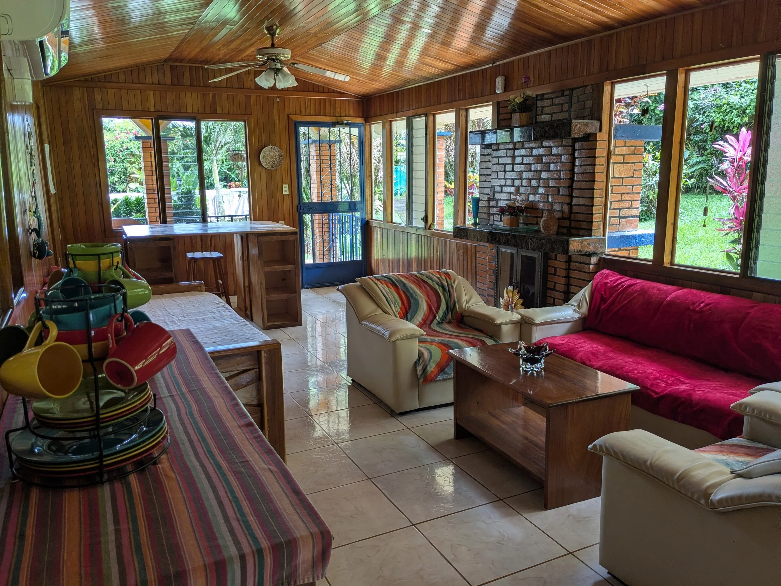 Living room with wooden walls and ceiling, tile floor, brick fireplace, red and cream sofas, coffee table, and large windows overlooking a green garden outside.