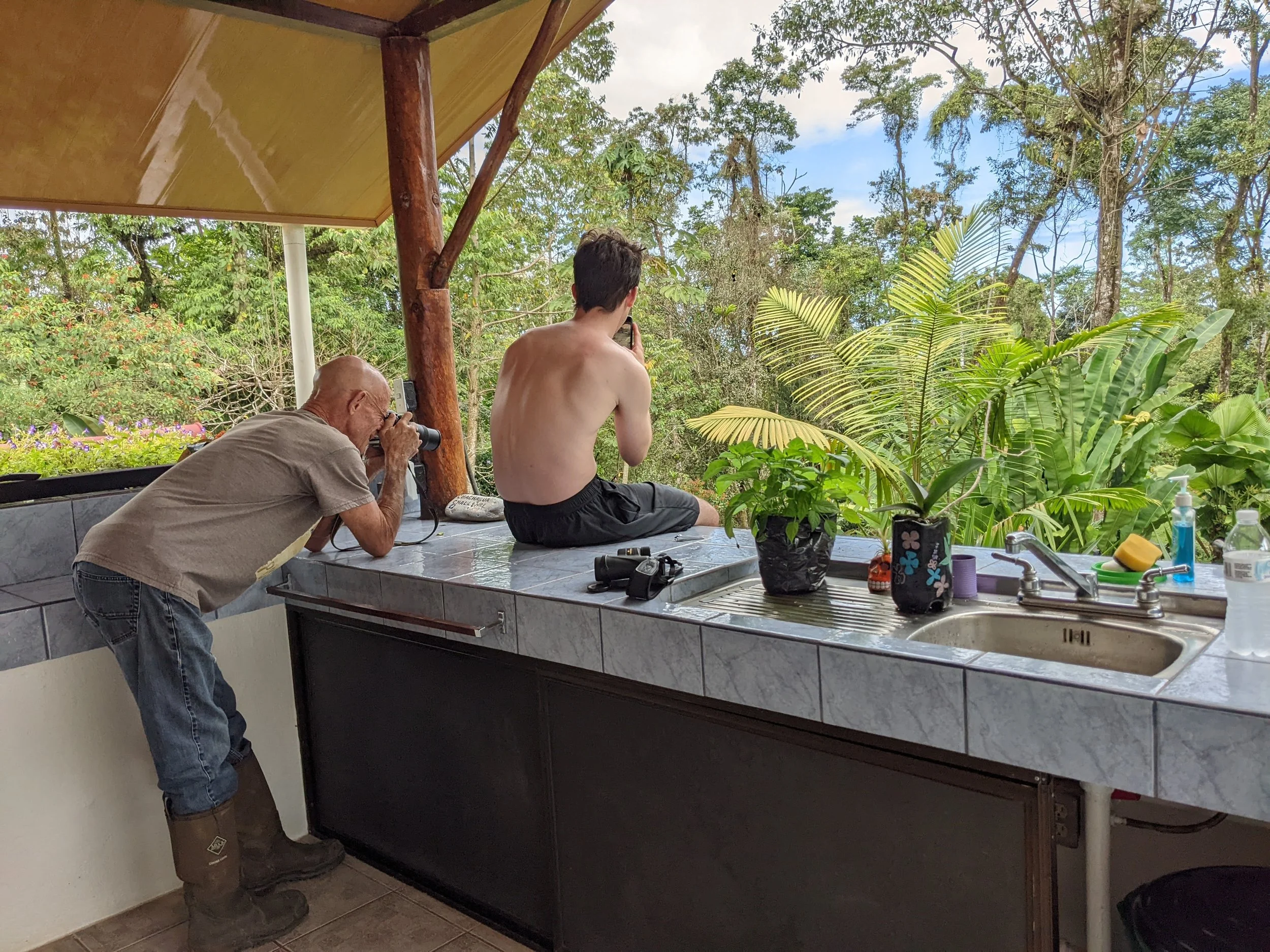 An elderly man taking a photograph of a shirtless young man sitting on a kitchen counter, with lush greenery in the background.