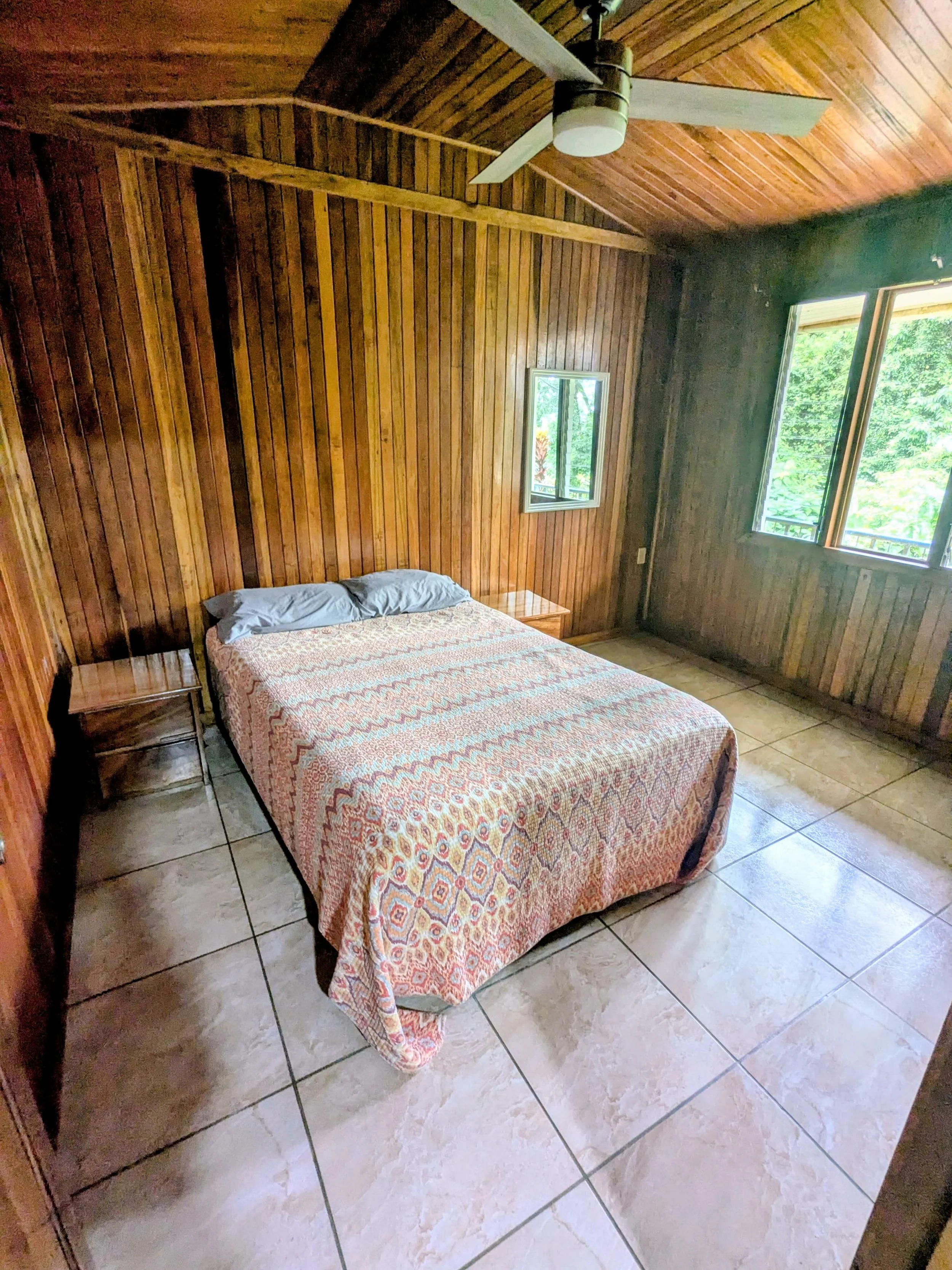 A cozy bedroom with wooden paneled walls, a bed with patterned bedspread, two nightstands, a small window, a ceiling fan, and large tiled floor.