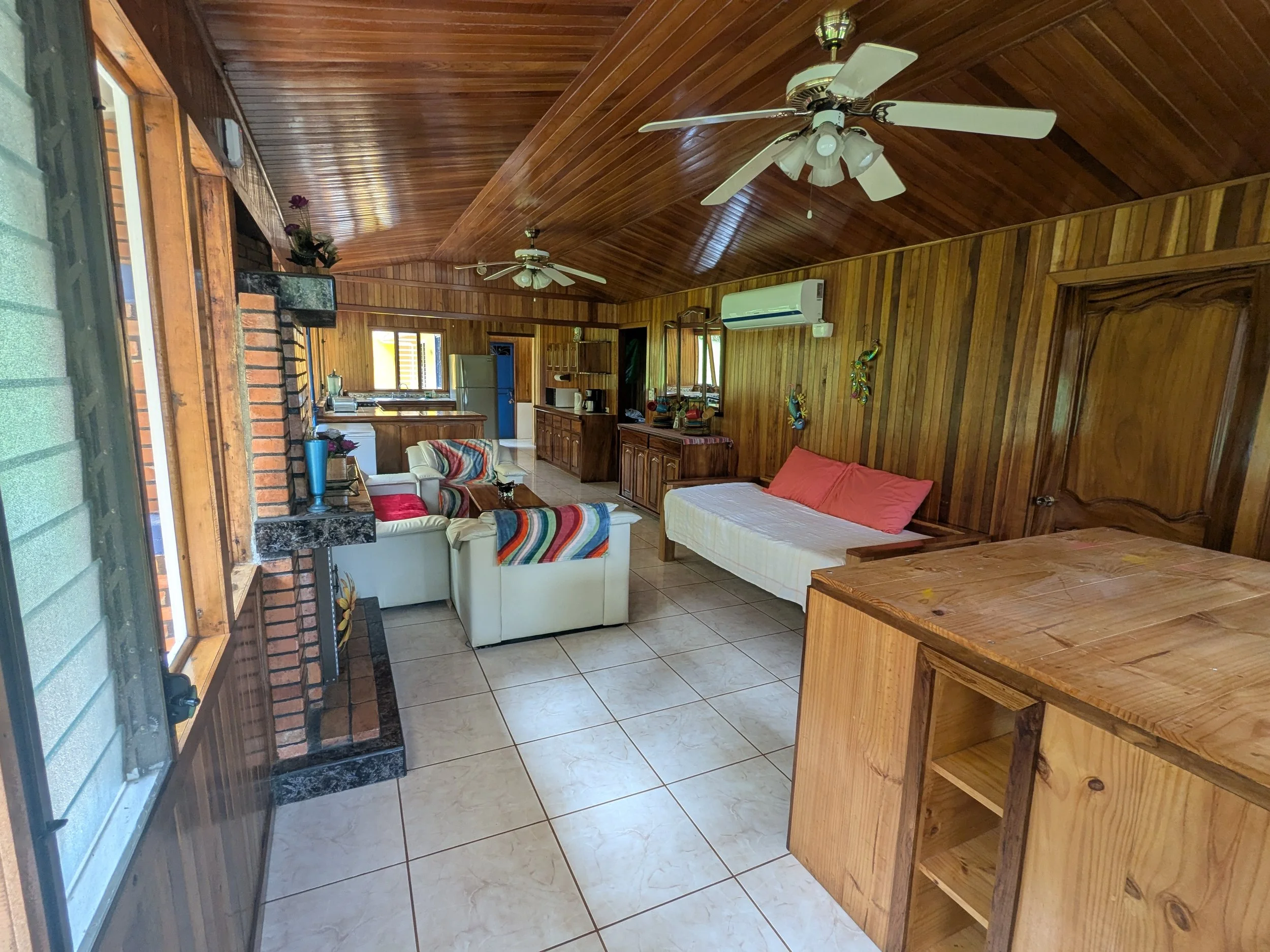 Living room with wood-paneled walls and ceiling, tiled floor, two ceiling fans, a couch, a daybed, a wooden cabinet, a fireplace, walls decorated with colorful art, a window, and kitchen in the background.