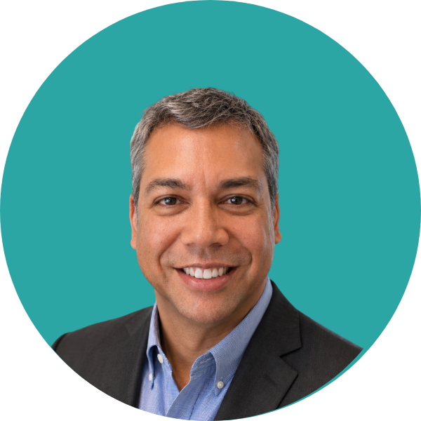 A smiling man with short gray hair, wearing a dark suit jacket and a light blue collared shirt, against a teal background.
