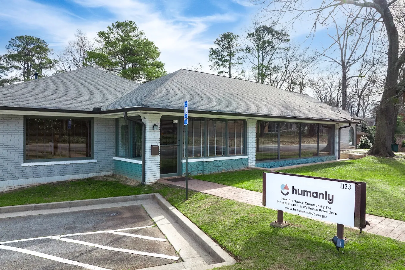 A white brick building with large windows, a small parking lot, and a sign that reads 'humanly, flexible space community for mental health & wellness providers, 1123' with a website URL. The building is surrounded by green grass and trees, with a clear blue sky above.