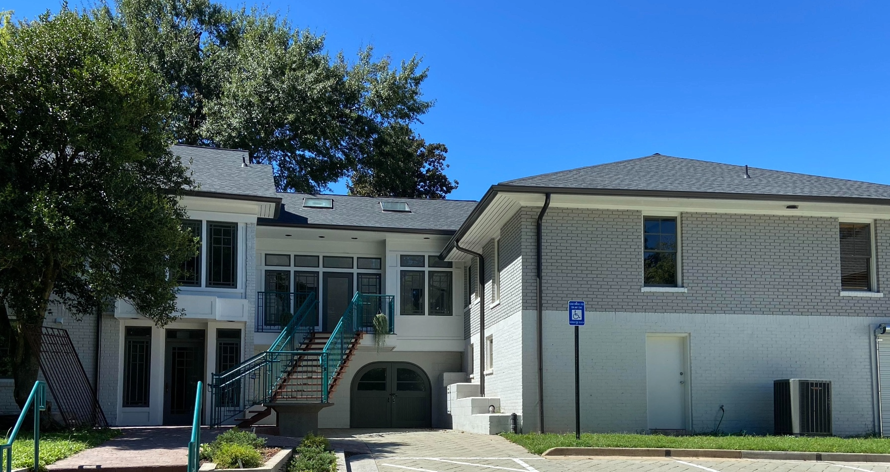 The back of a white brick residential building with a staircase, a handicap parking sign, windows, an air conditioning unit, and a tree in the foreground.