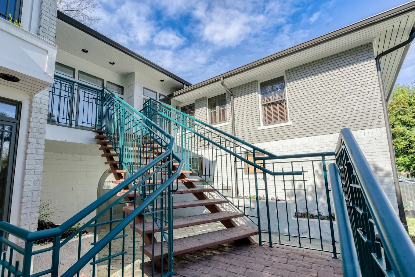 Exterior view of a modern house with white brick walls, large windows, and a teal metal staircase leading to an upper balcony.