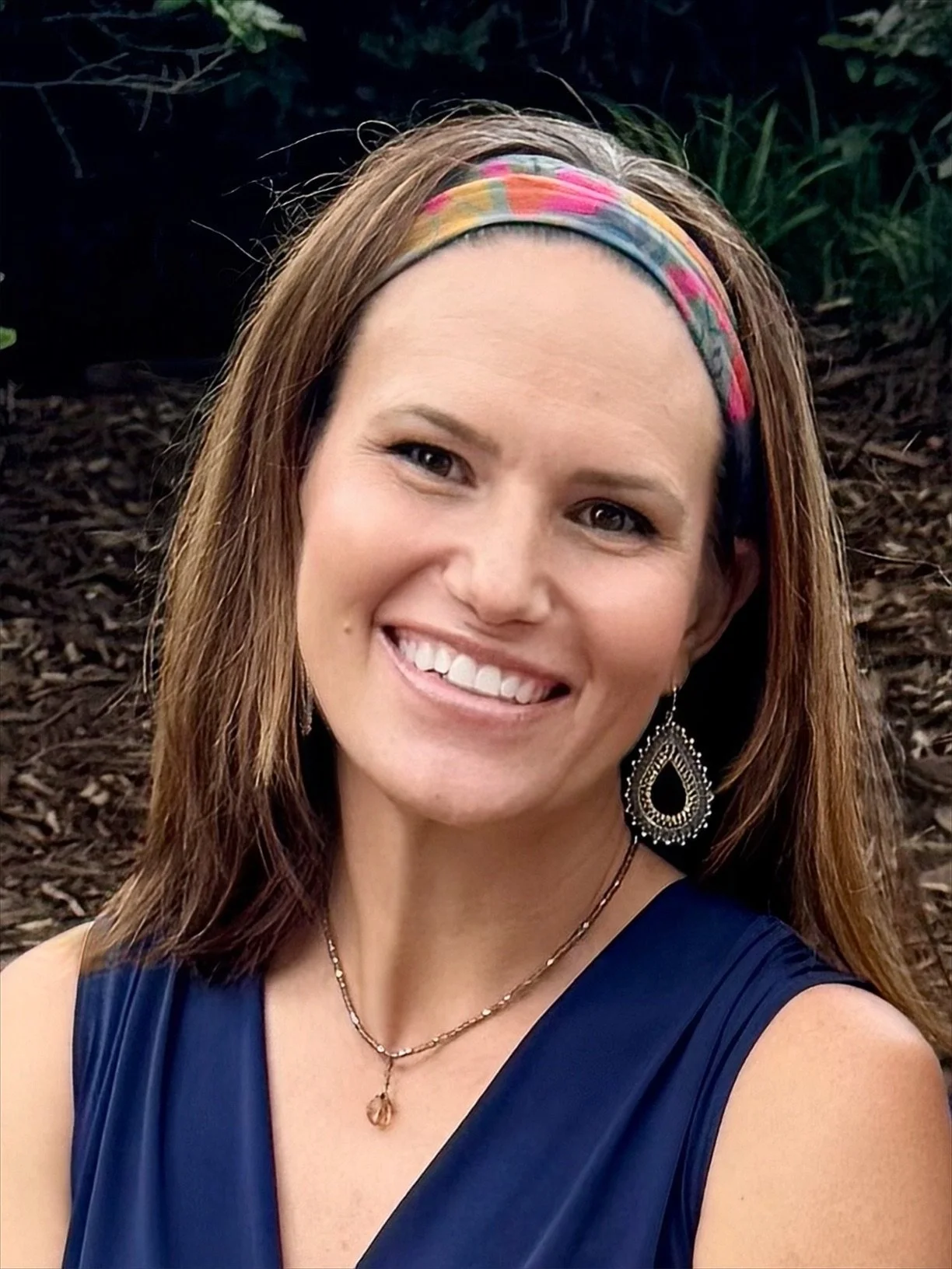 A woman with brown hair wearing a colorful headband, earrings, a necklace, and a blue dress, smiling outdoors with dark soil and greenery in the background.