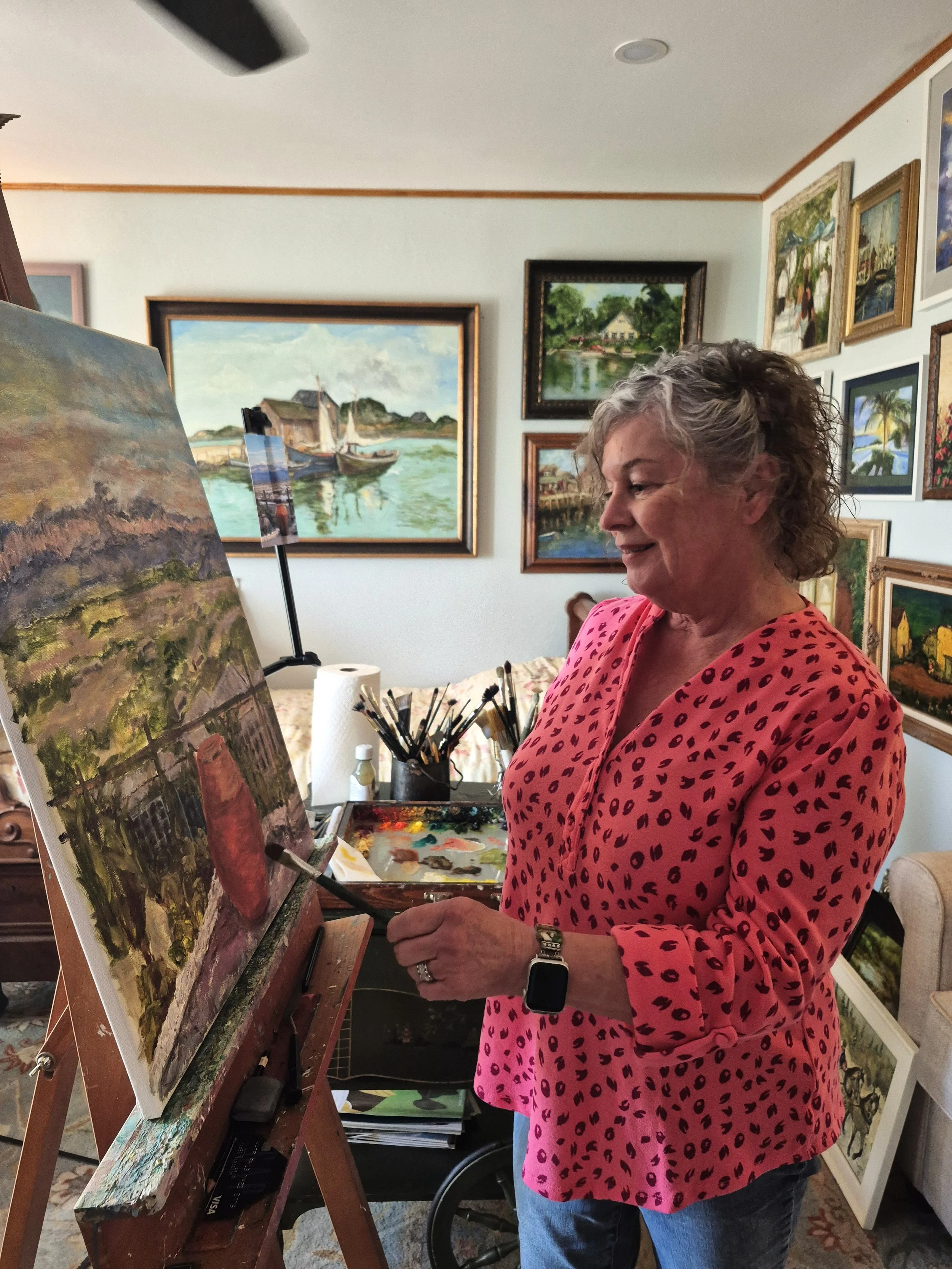A woman with girly hair paints a picture at an eisel in a well lit room surrounded by other paintings she has done.