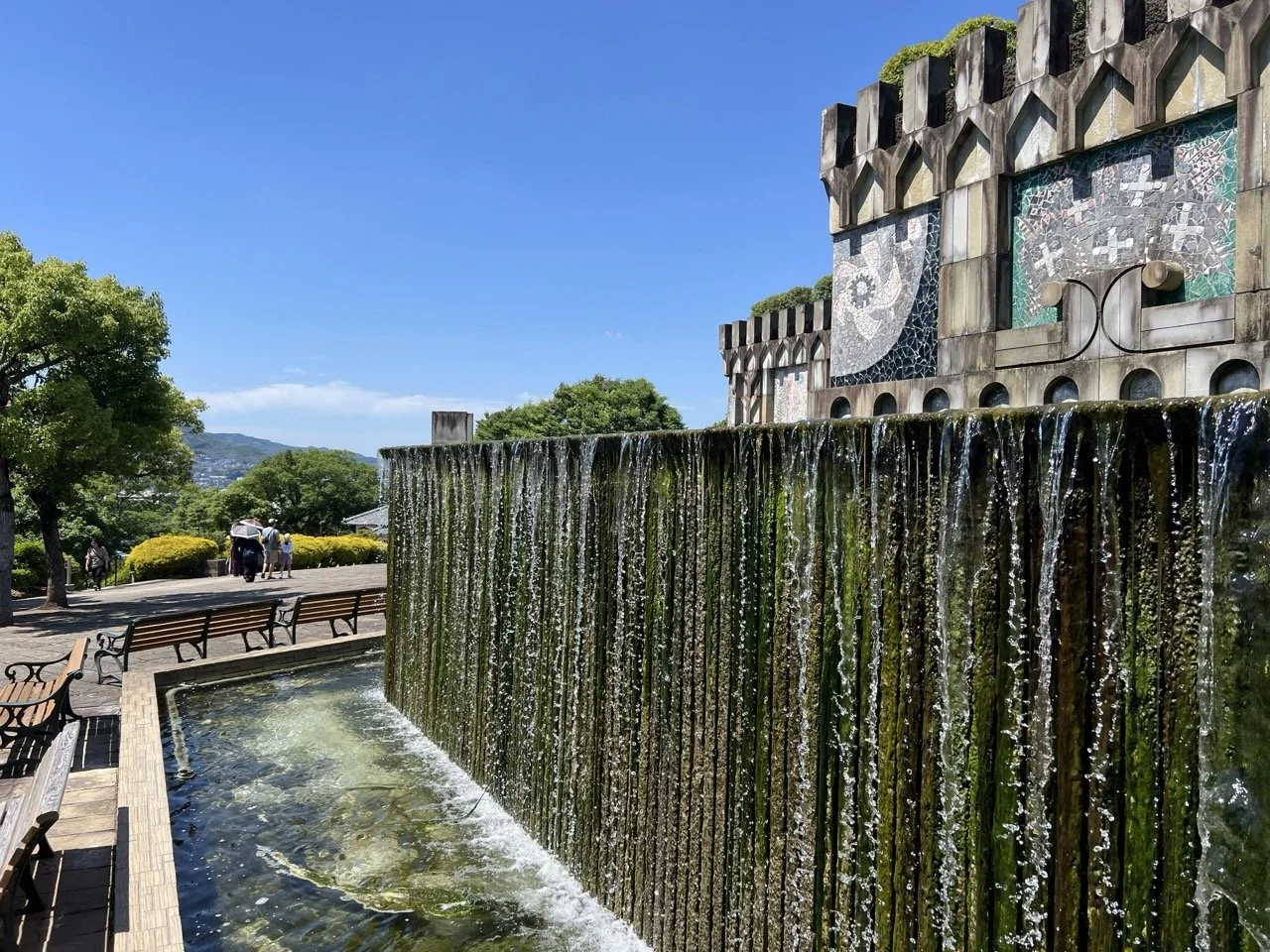 A long wall adorned with fountains and colorful tiles sits in a garden on a hillside over a city below.