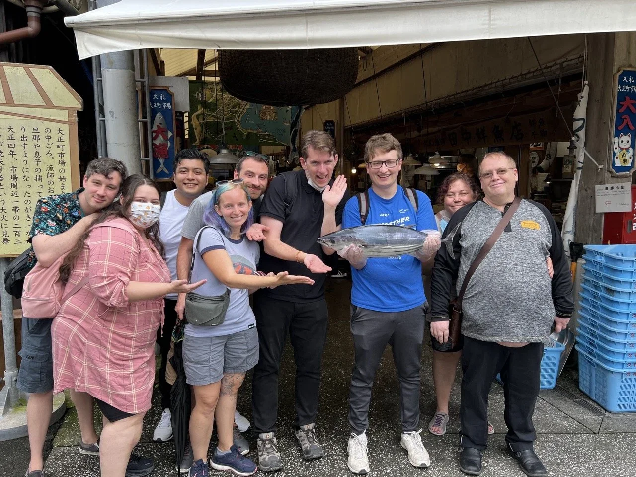 A group of twelve people posing for a photo outdoors, one person holding a large fish, some wearing masks, in front of a market or shop with signage and maps.