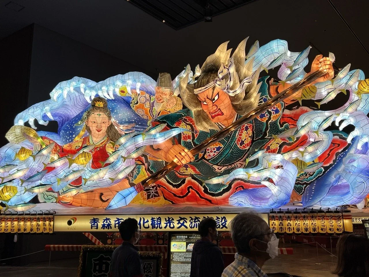 Large illuminated lantern display depicting fierce warrior and a woman surrounded by swirling patterns and clouds, with people observing in front.
